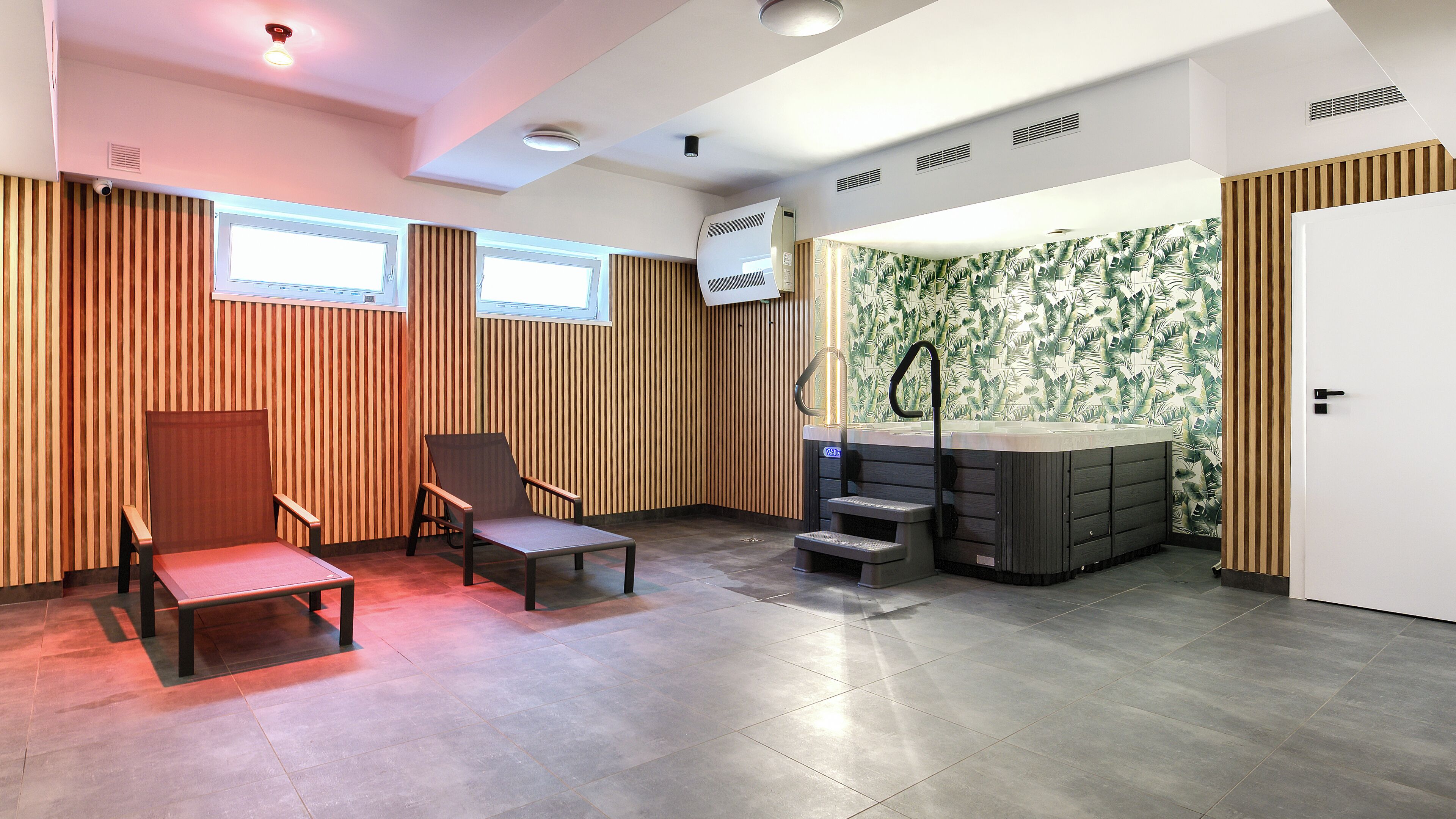 A modern room with a wooden slatted wall, a seating area with a bench and chairs, a hot tub, and a door with a patterned wallpaper.