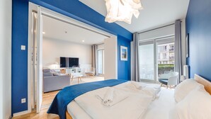 A modern hotel room with a large bed, a TV, a seating area with a sofa and table, and a view of the outside.