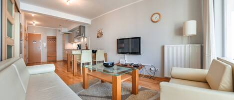 Ein modernes Wohnzimmer mit Glastisch, weißen Sofas, einem Flachbildfernseher und einer Küche im Hintergrund.