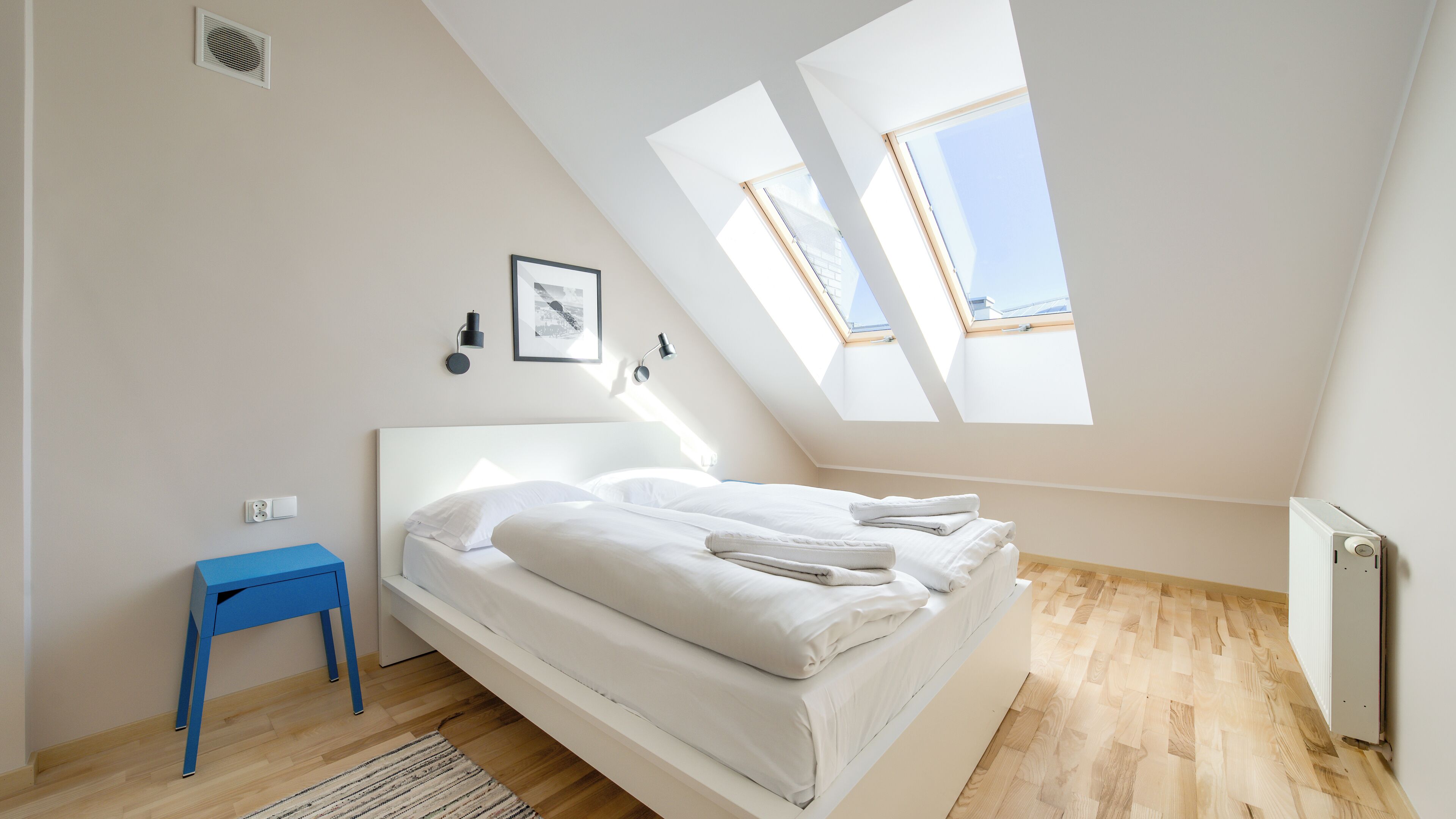 Une chambre avec un plafond en pente, des fenêtres de toit, un lit, une petite table et un radiateur.