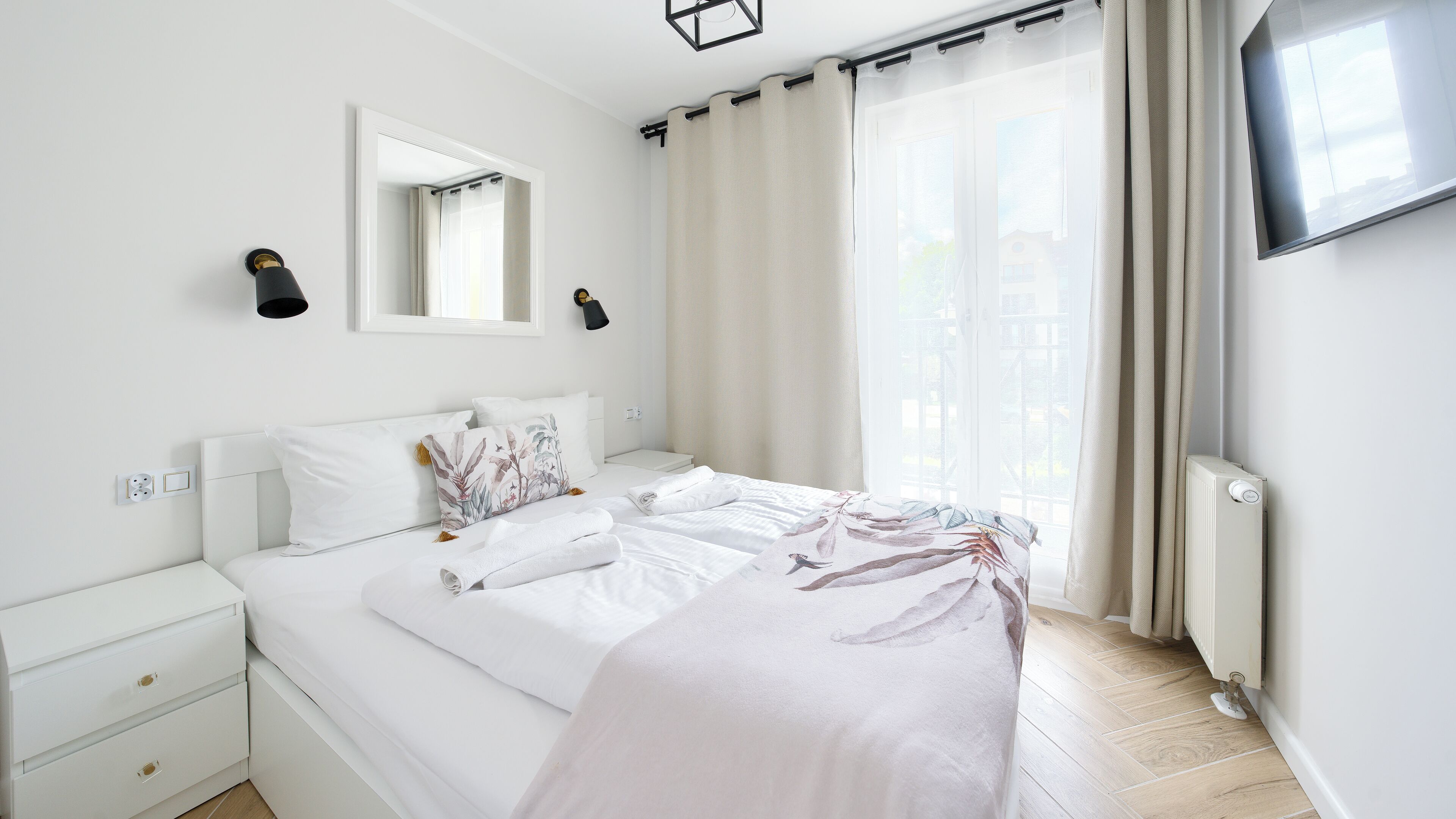 A modern bedroom with a large bed, a bedside table, a mirror, and a TV on the wall.