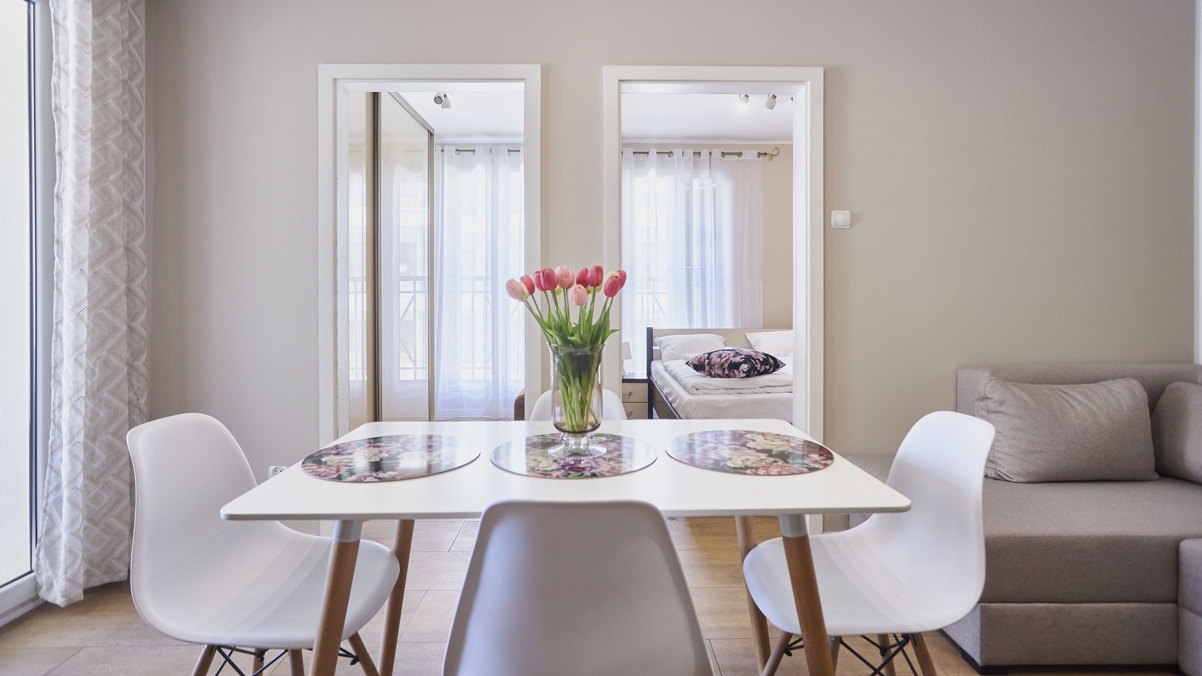 Un comedor con una mesa blanca, dos sillas blancas y un jarrón de tulipanes rosados. En el fondo, un sofá color crema.