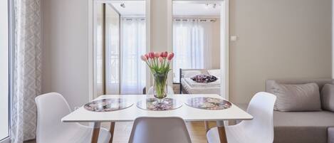 Un comedor con una mesa blanca, dos sillas blancas y un jarrón de tulipanes rosados. En el fondo, un sofá color crema.
