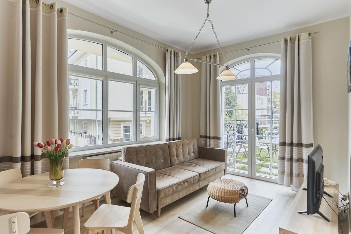 Un salon spacieux avec un canapé, une table ronde et des chaises. On y trouve un téléviseur posé sur un meuble et une fenêtre avec des rideaux.