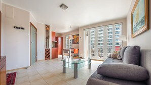 A spacious living area with a glass coffee table, a grey sofa, and a view of a high-rise building through large windows. - Apartamenty Sun & Snow Promenada (Swinoujscie)