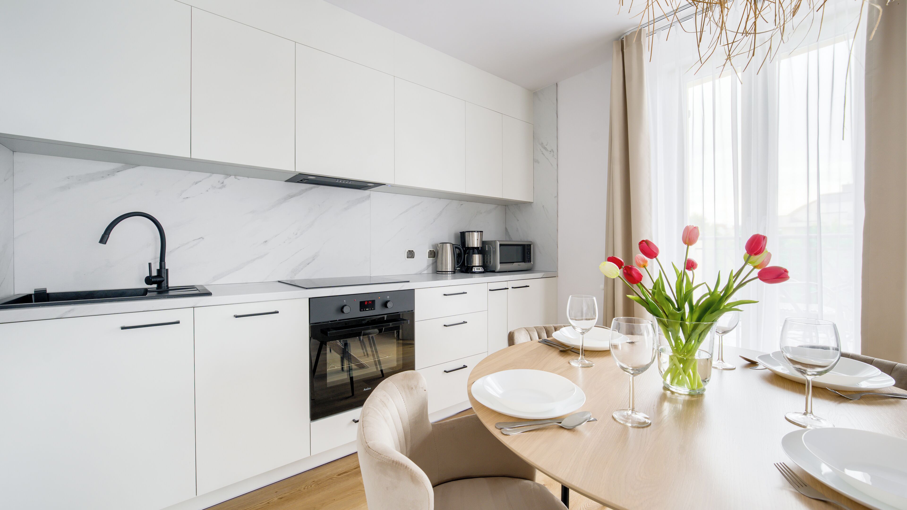 Une cuisine moderne avec des meubles blancs, un robinet noir et un coin repas avec une table dressée pour deux personnes.