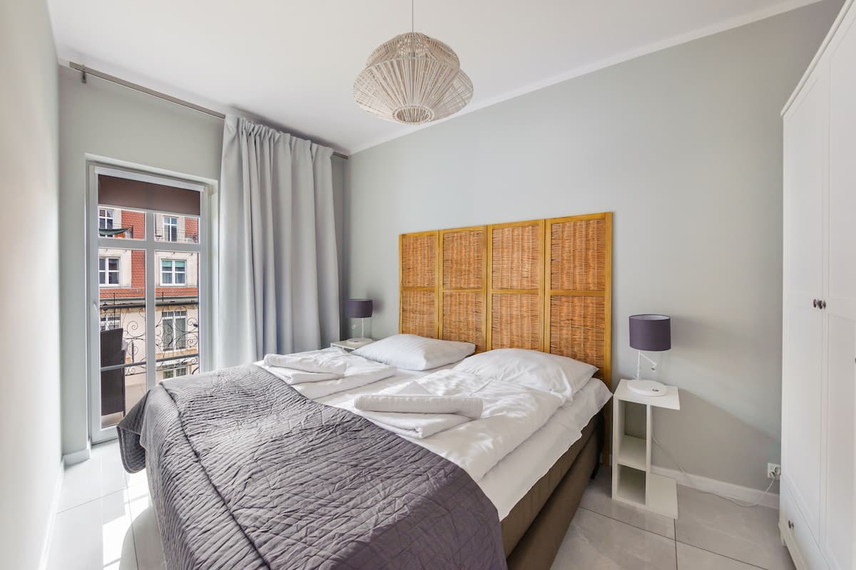 Une chambre moderne avec un grand lit, une table de chevet avec une lampe, une tête de lit en osier et une vue sur la ville par une grande fenêtre.
