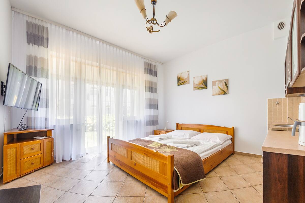 Une chambre d’hôtel avec un lit en bois, un bureau doté d’un téléviseur à écran plat, une machine à café et une grande fenêtre ornée de rideaux légers.