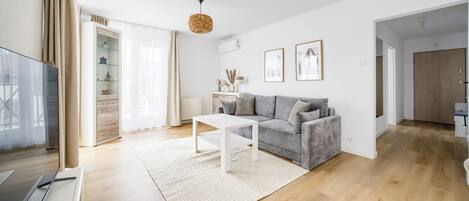A modern living room with a grey sofa, white coffee table, and a large window with sheer curtains.