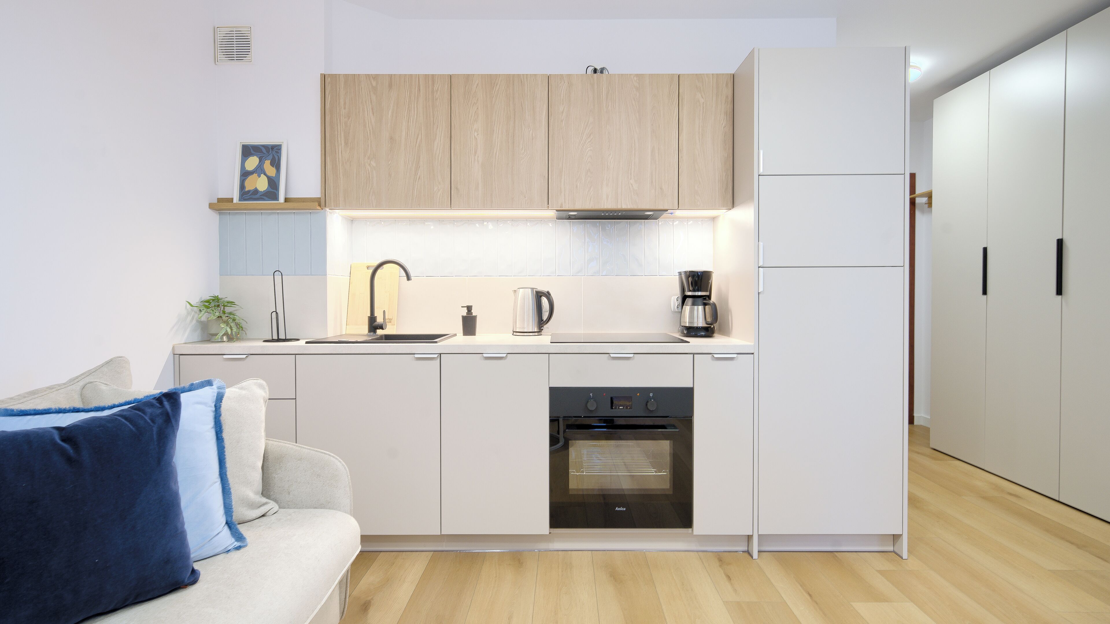A modern kitchen with white cabinets, a stainless steel sink, a kettle, a coffee maker, and a built-in oven.