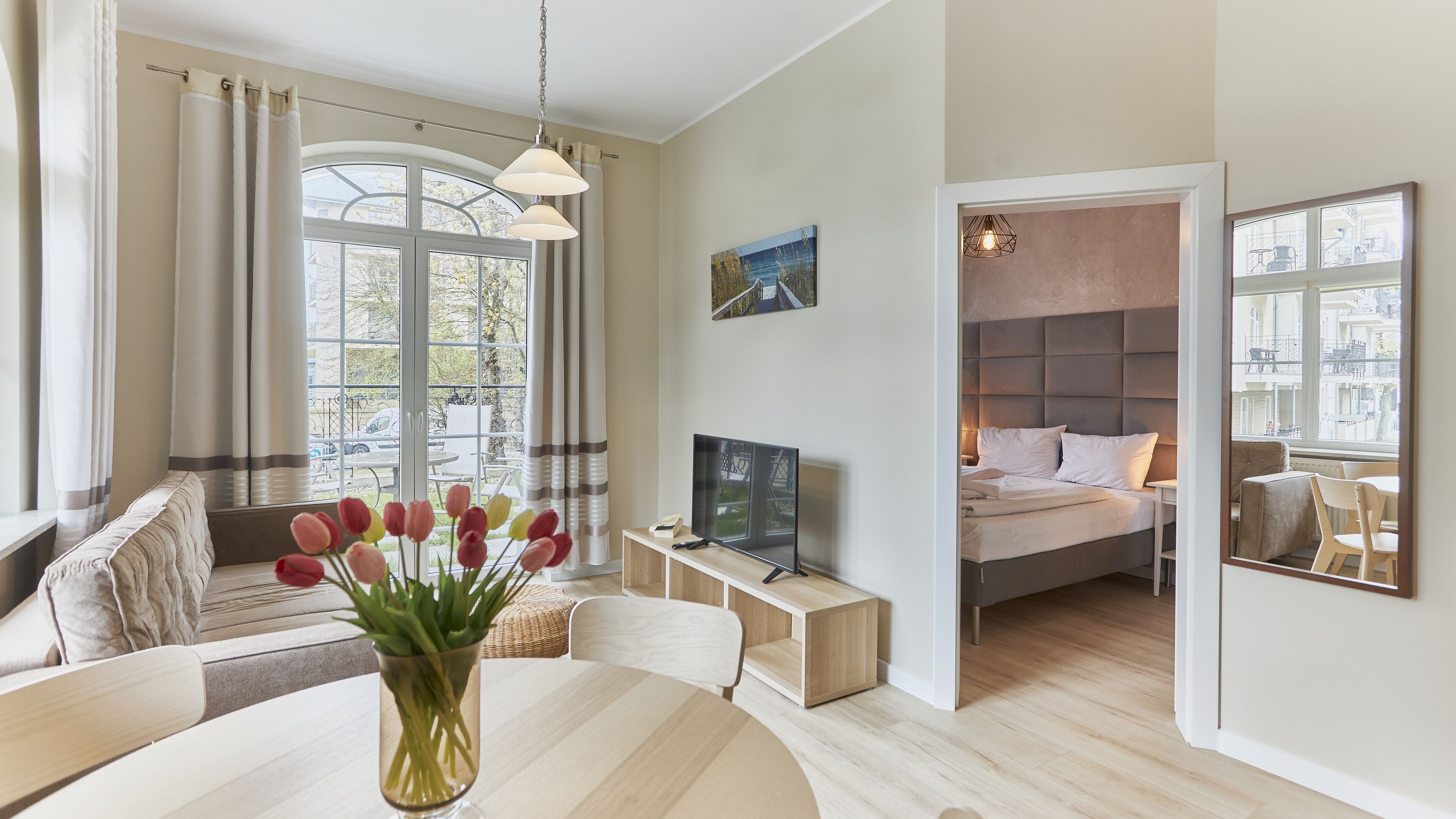 A modern living room with a sofa, a dining table with a vase of tulips, a TV on a wooden stand, and a bed with a view of the city.