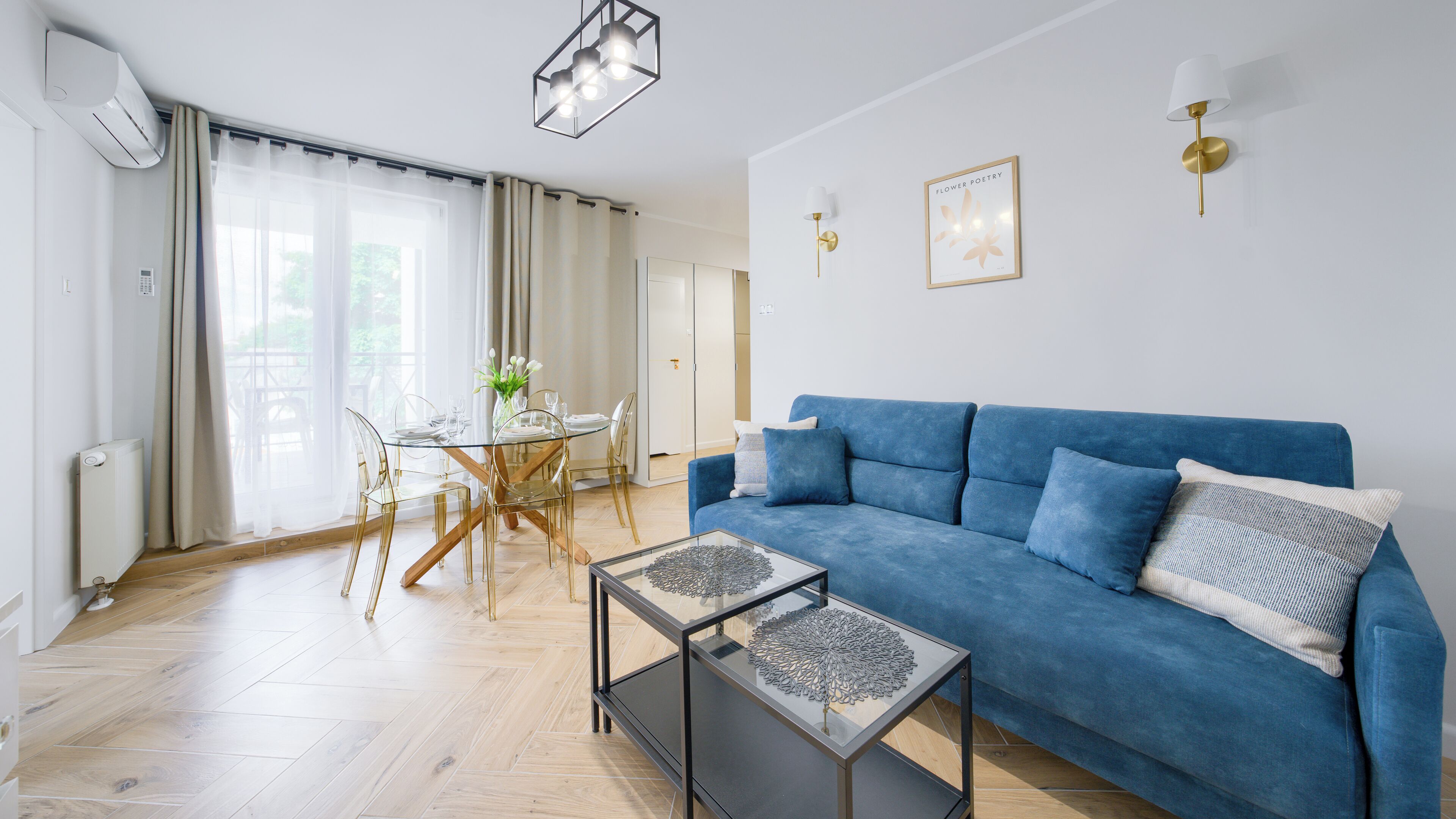 A modern living room with a blue sofa, glass coffee table, and a dining area with a glass table and chairs.