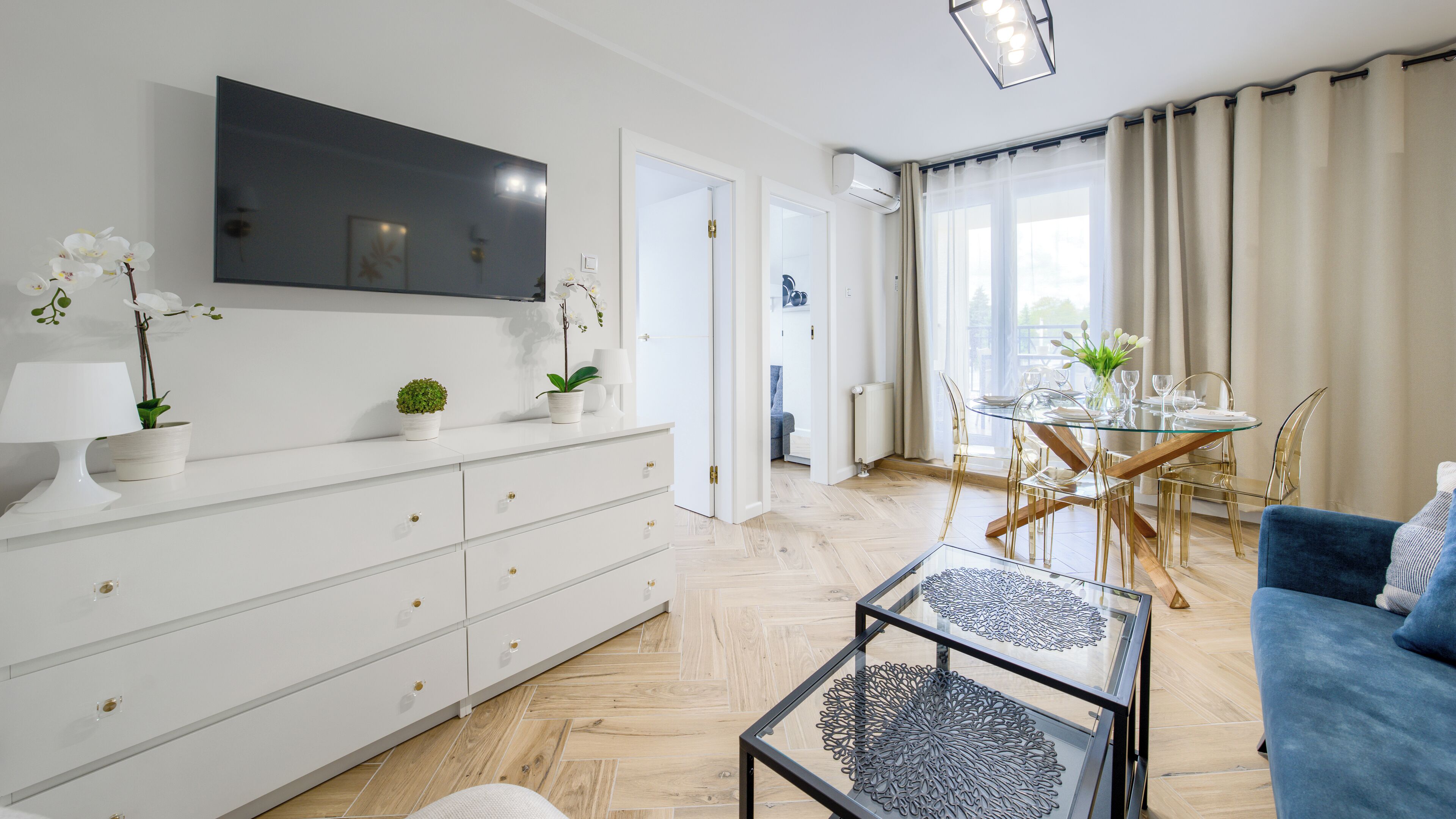 Uma sala de estar moderna com televisão de tela plana, armários brancos, uma mesa de centro de vidro e um sofá azul.