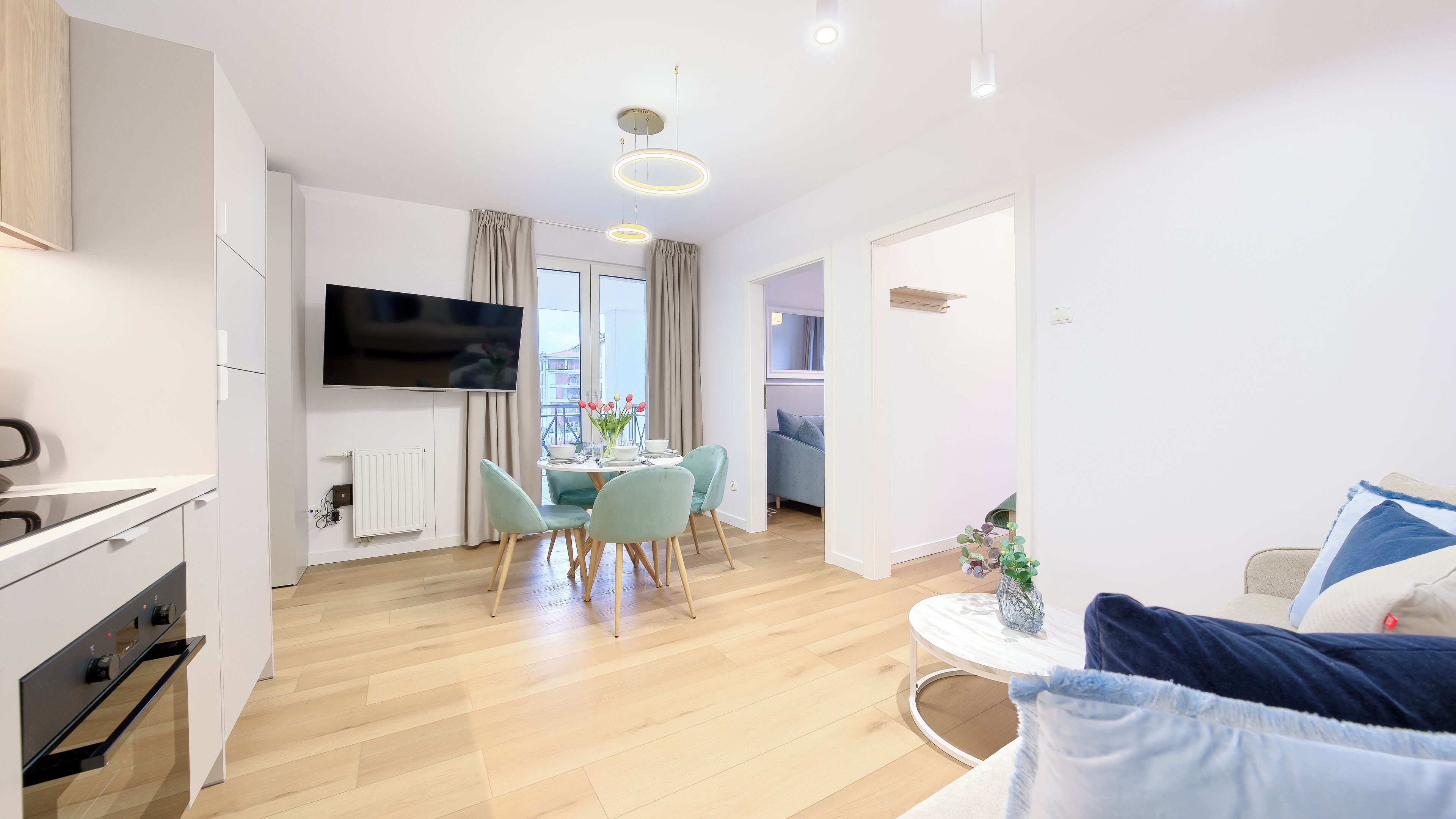 Un appartement moderne comprenant une cuisine, un coin repas et un salon. Il est équipé d’un téléviseur à écran plat, d’une table à manger avec des chaises, d’un canapé et d’une petite table d’appoint agrémentée d’une plante.