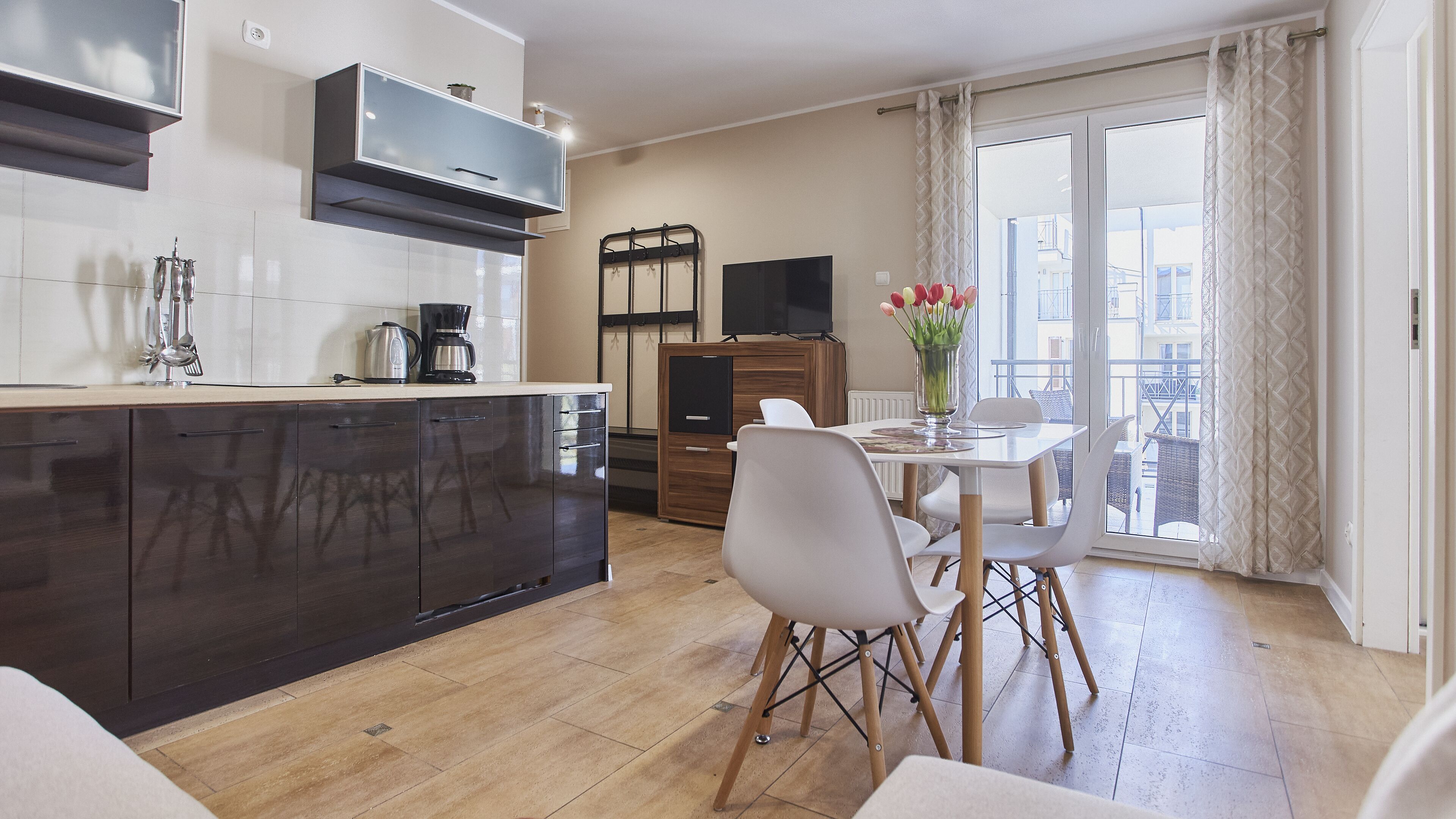 Een moderne keuken met donkere kasten, een koffiezetapparaat en een televisie boven de aanrecht. Er is een zithoek met een tafel en stoelen, en een balkon met uitzicht op de stad.