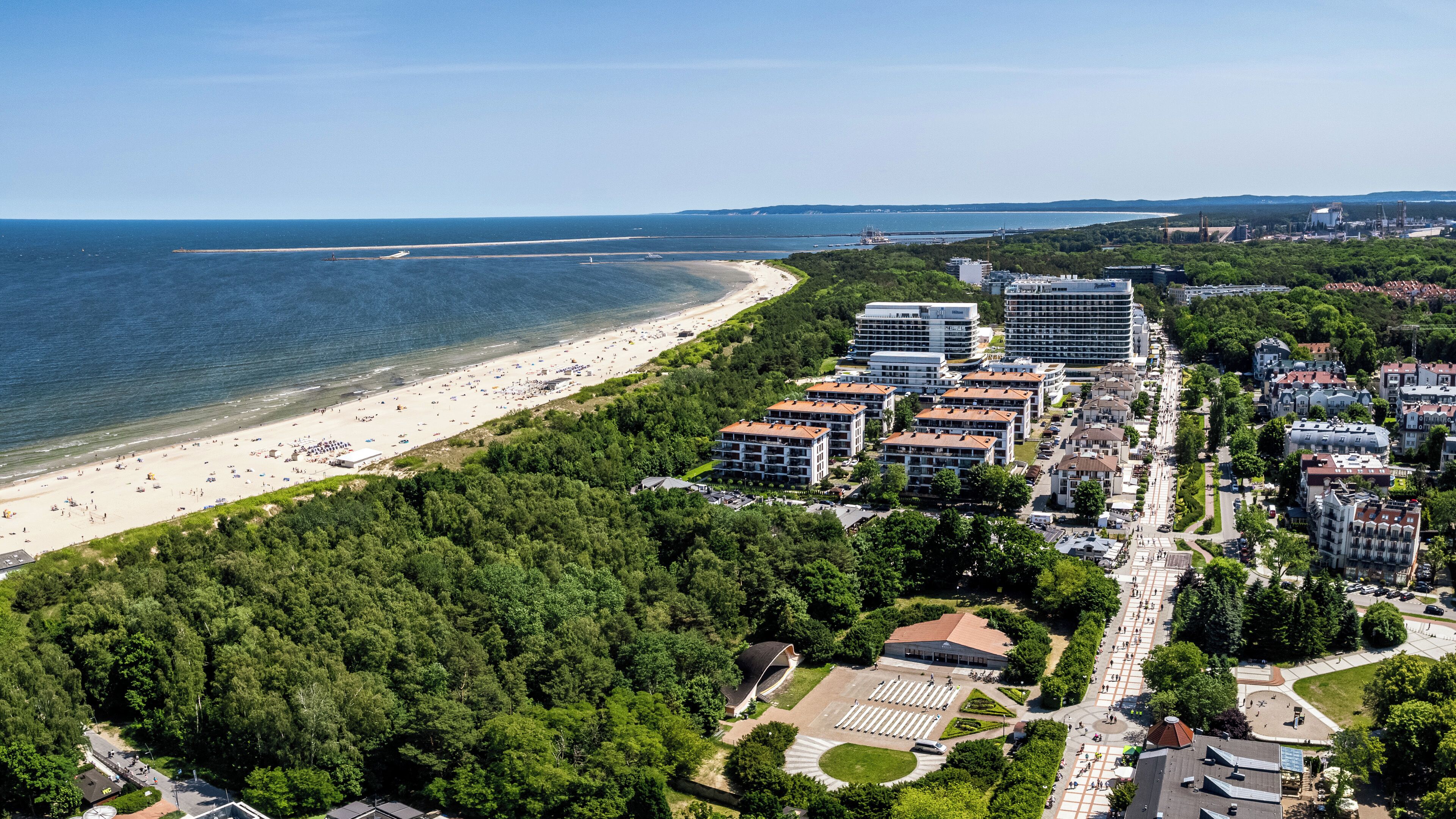 An aerial view of a coastal area with a beach, dense greenery, and modern buildings.