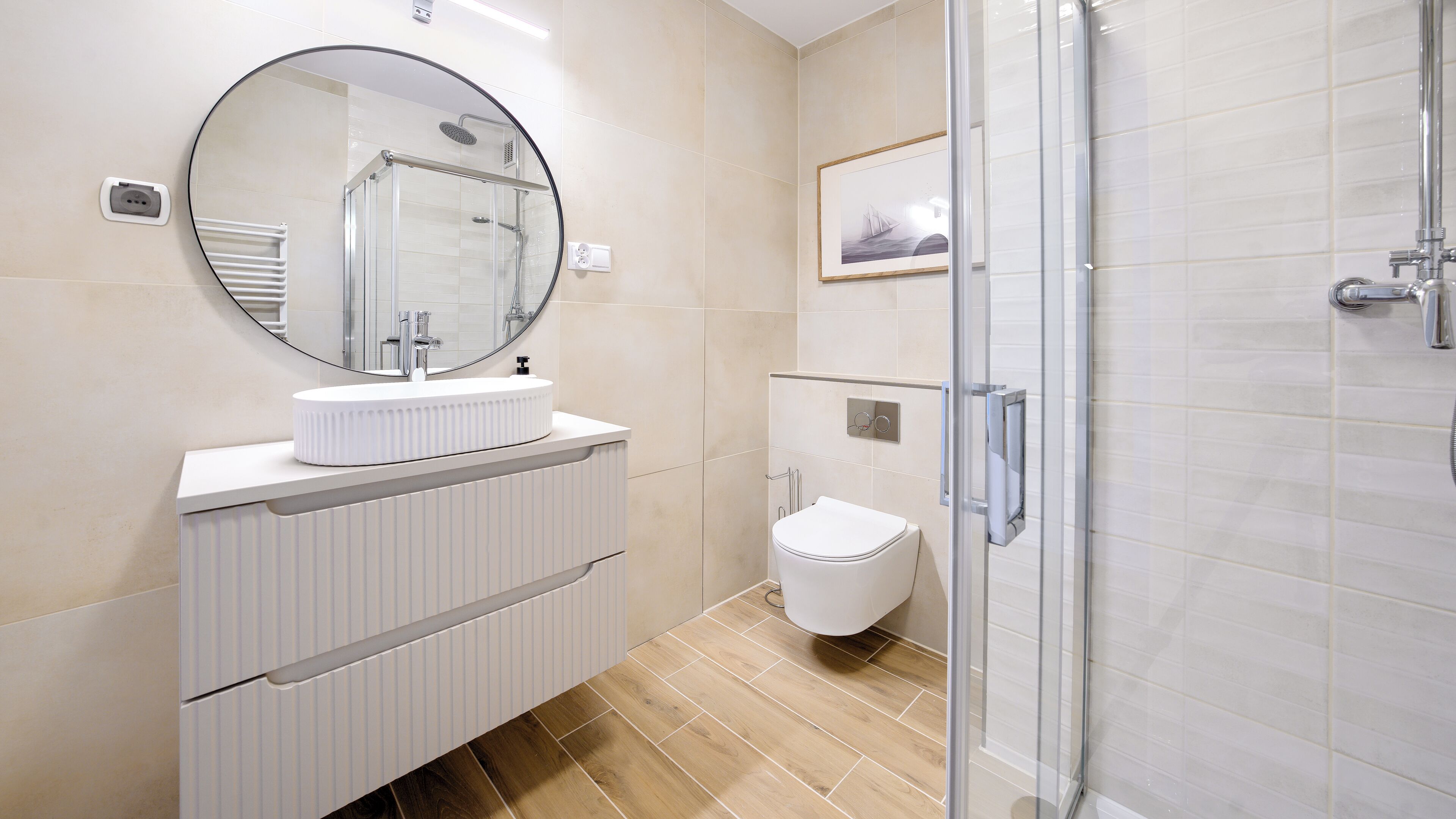Une salle de bain moderne avec un grand miroir rond, un lavabo blanc, des toilettes et une douche séparée par une paroi de verre.