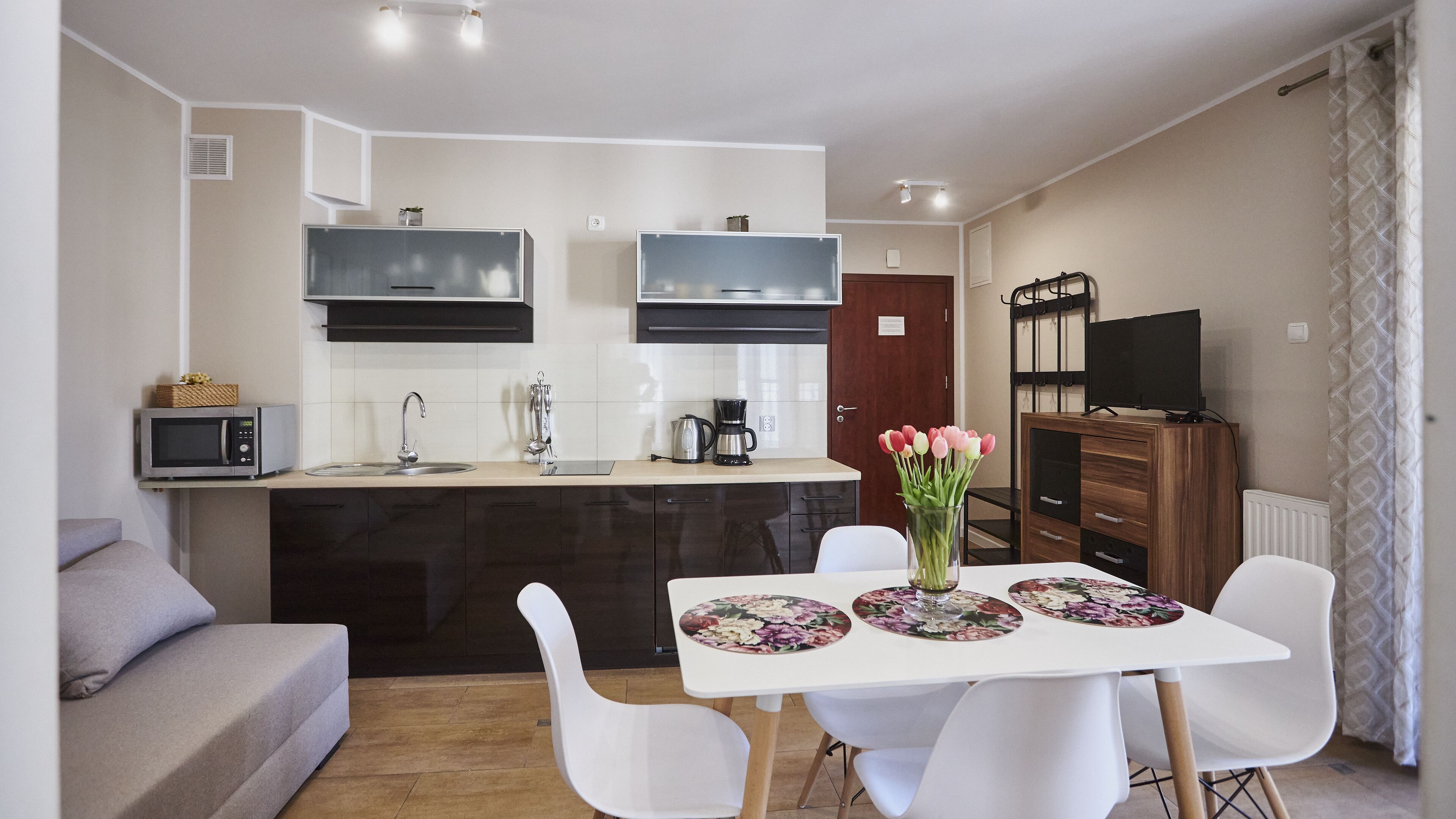 Cozinha moderna com micro-ondas embutido, máquina de café e chaleira. Há uma área de jantar com mesa e cadeiras brancas, e um vaso de flores sobre a mesa.