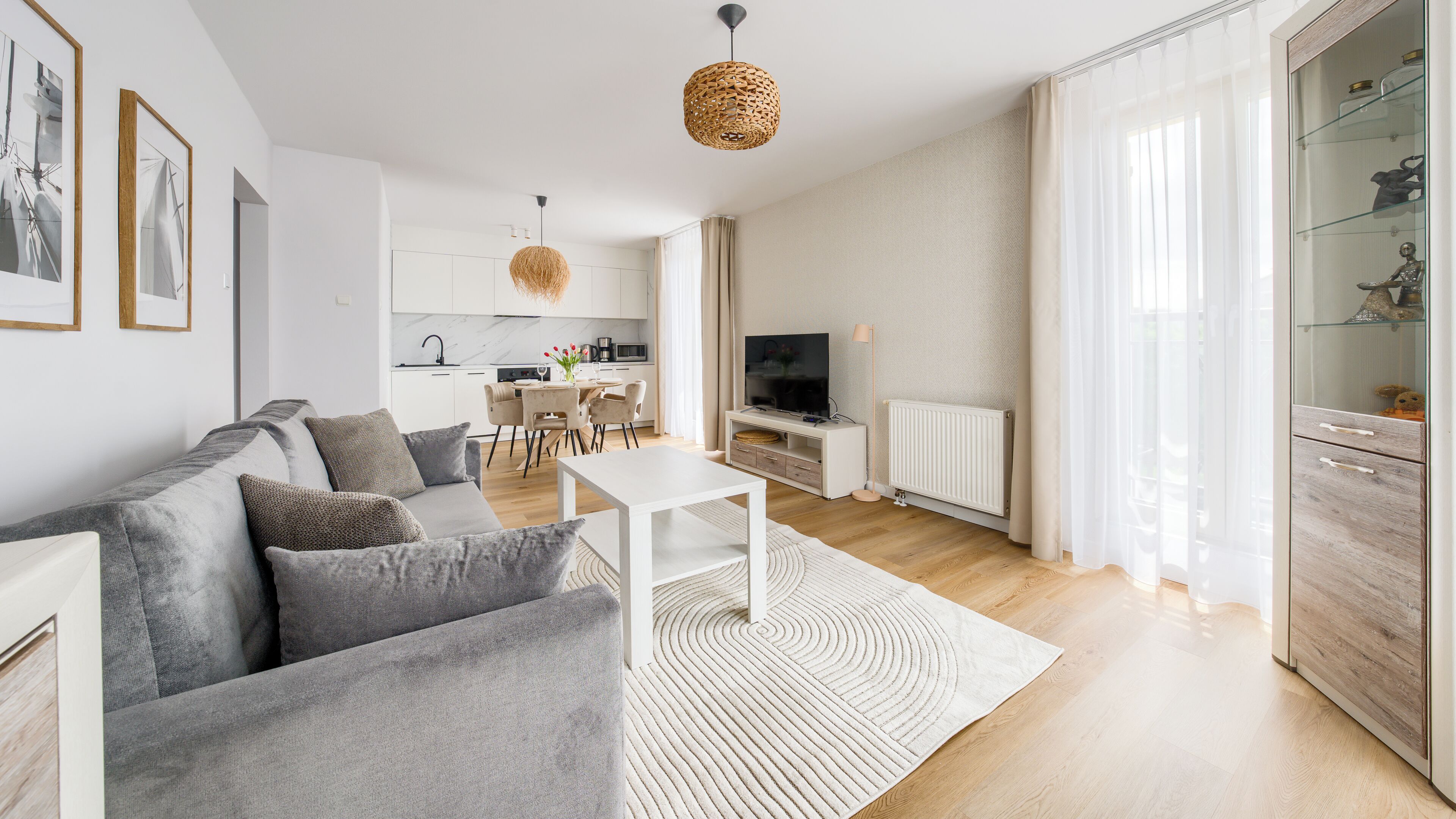 Een moderne woonkamer met een grijze bank, een witte salontafel en een televisie. In de achtergrond bevindt zich een keuken met witte kasten en een eethoek.