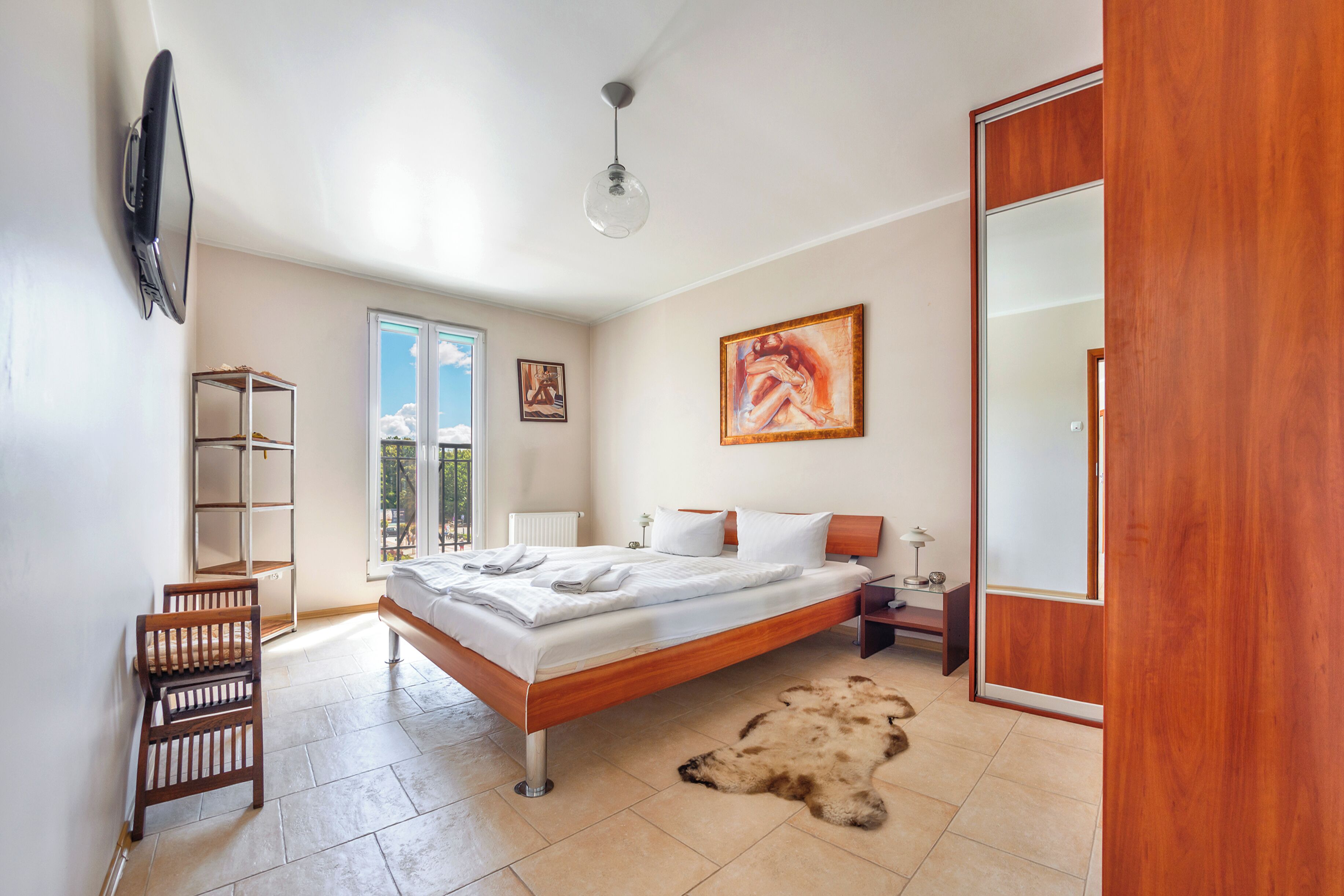 A bedroom with a bed, a wooden wardrobe, a mirror, a painting, and a view of the outdoors.