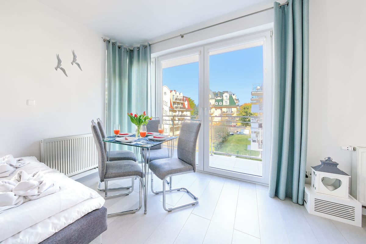 Une chambre d’hôtel moderne équipée d’un lit, d’une table à manger avec des chaises, offrant une vue sur la ville grâce à de grandes fenêtres, et dotée d’un balcon avec vue sur le quartier.
