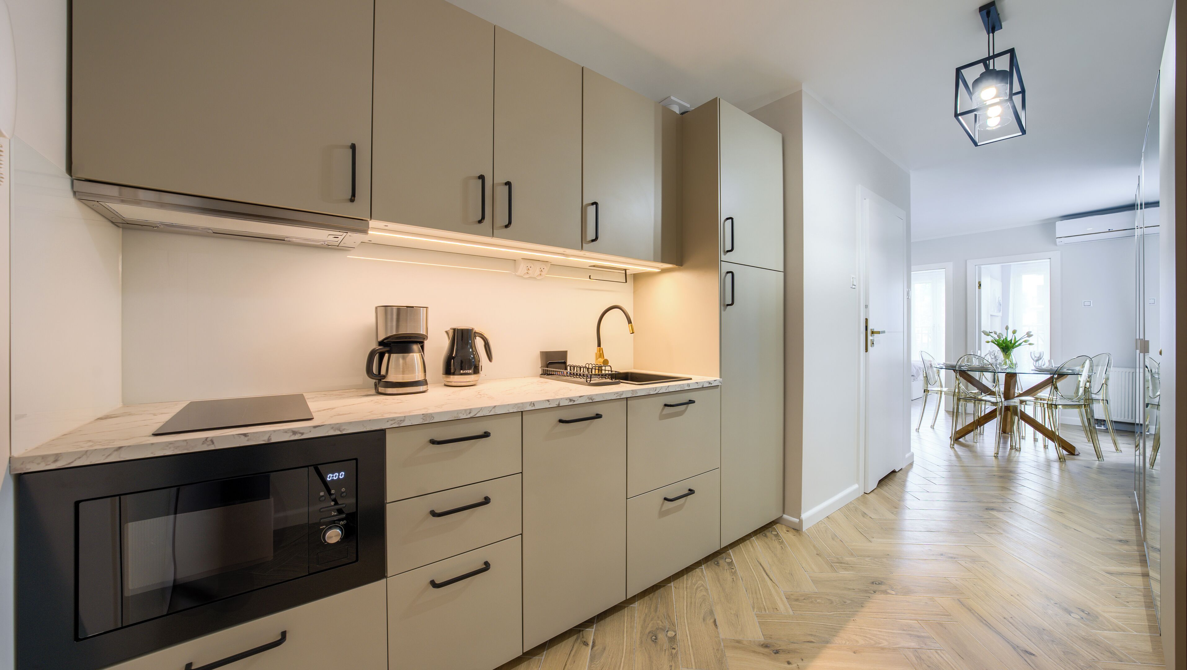 A modern kitchen with built-in appliances, a dining area with a glass table and chairs, and a hallway leading to a bedroom.