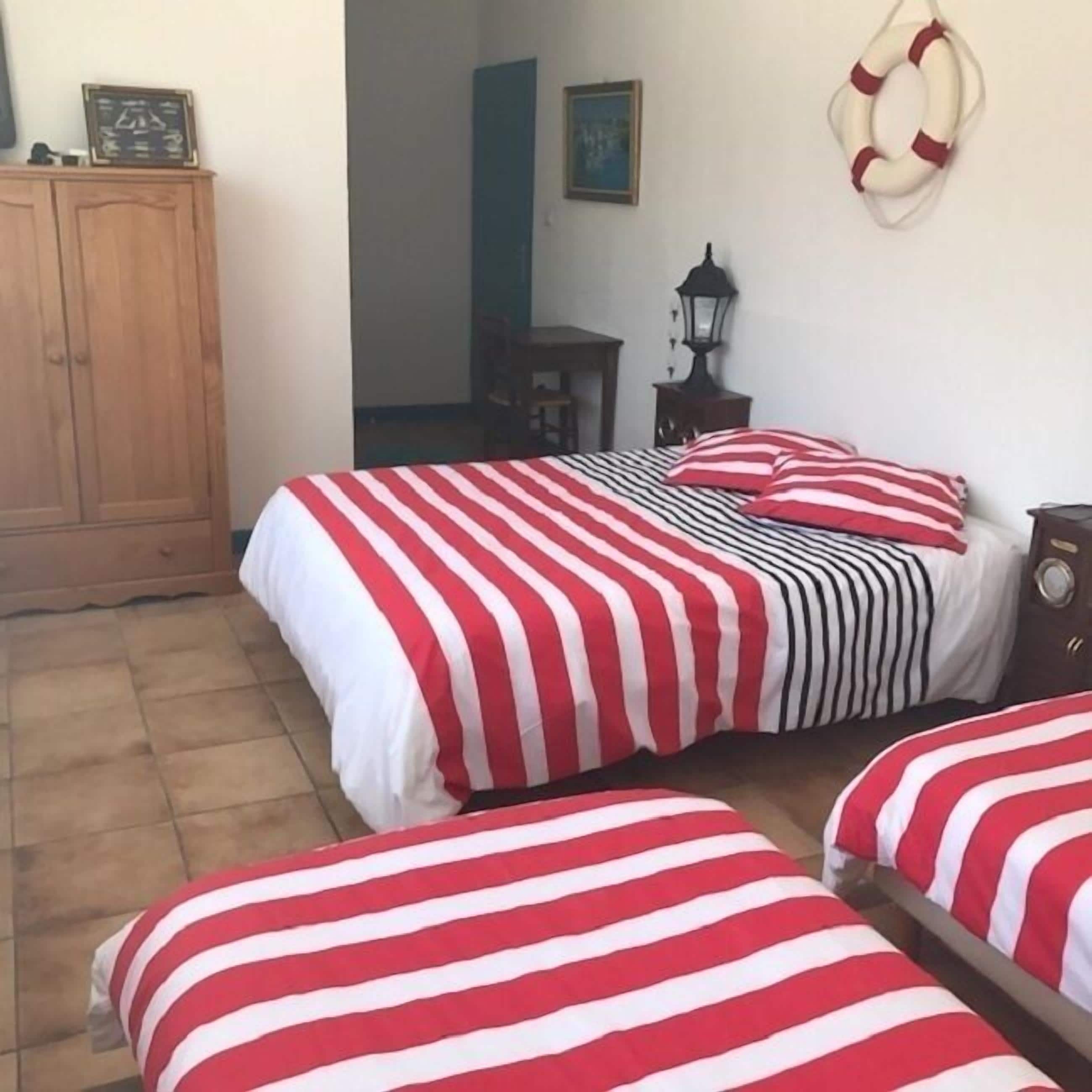 Une chambre avec deux lits, chacun recouvert d’une literie à rayures rouges et blanches. On y trouve une armoire en bois, une petite table avec une lampe et un gilet de sauvetage fixé au mur.