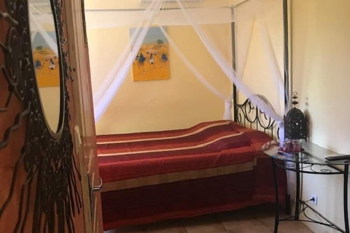 A bedroom with a four-poster bed, a bedside table with a lamp, and a wall-mounted air conditioner.