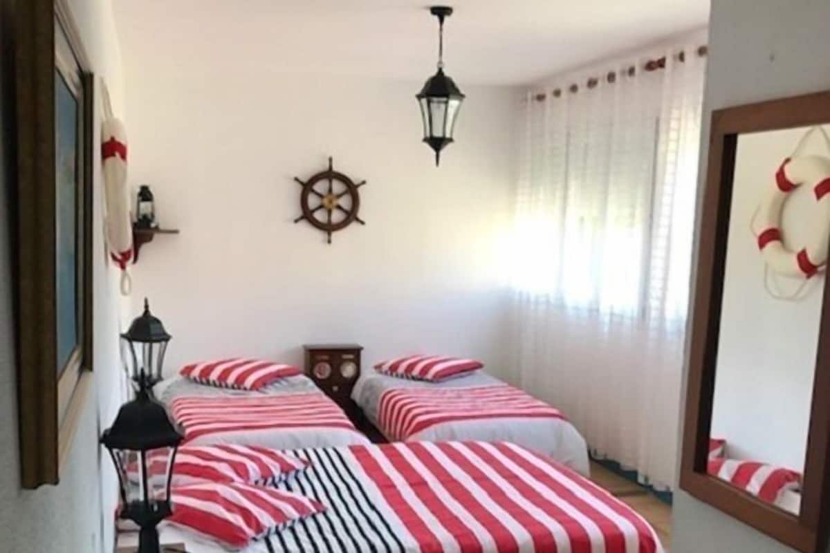 A room with two beds, each with red and white striped bedding, a wooden headboard, a wall-mounted ship's wheel, and two hanging lanterns.