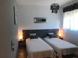 Habitación de hotel con dos camas, una lámpara de araña moderna, una obra de arte enmarcada en la pared y un ventanal con cortinas.