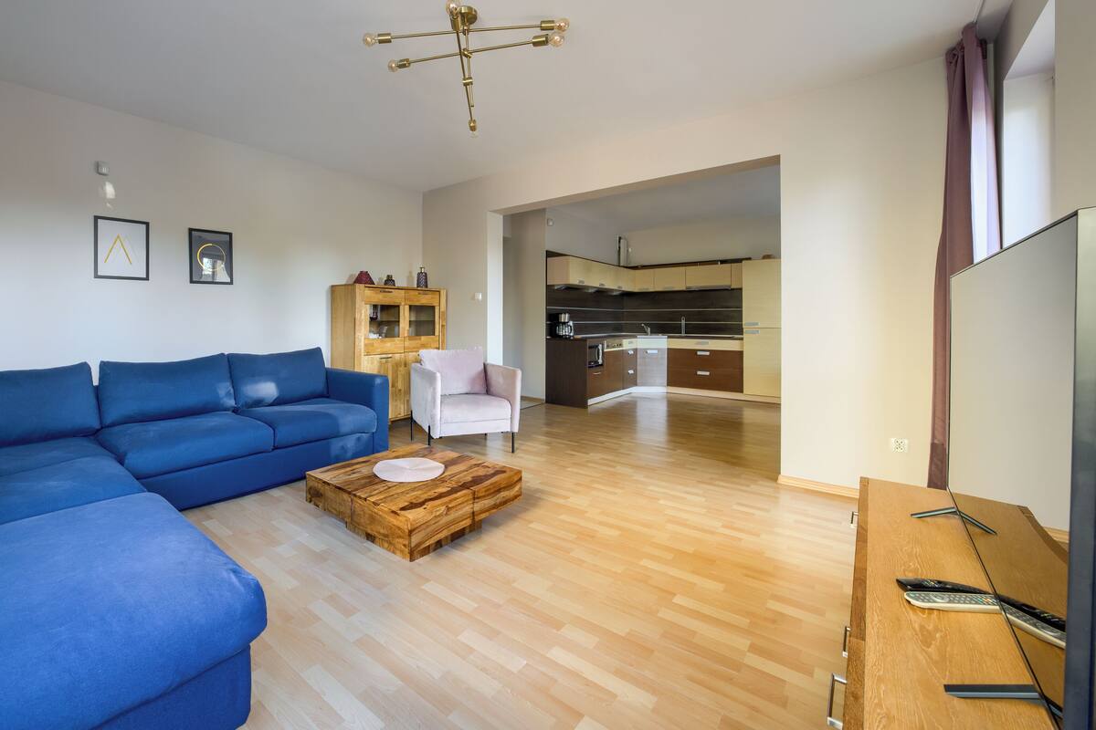 Un salon moderne avec un canapé d’angle bleu, une table basse en bois et un petit fauteuil. On aperçoit une cuisine à l’arrière-plan, avec des meubles de rangement en bois et une hotte de cuisinière en acier inoxydable.