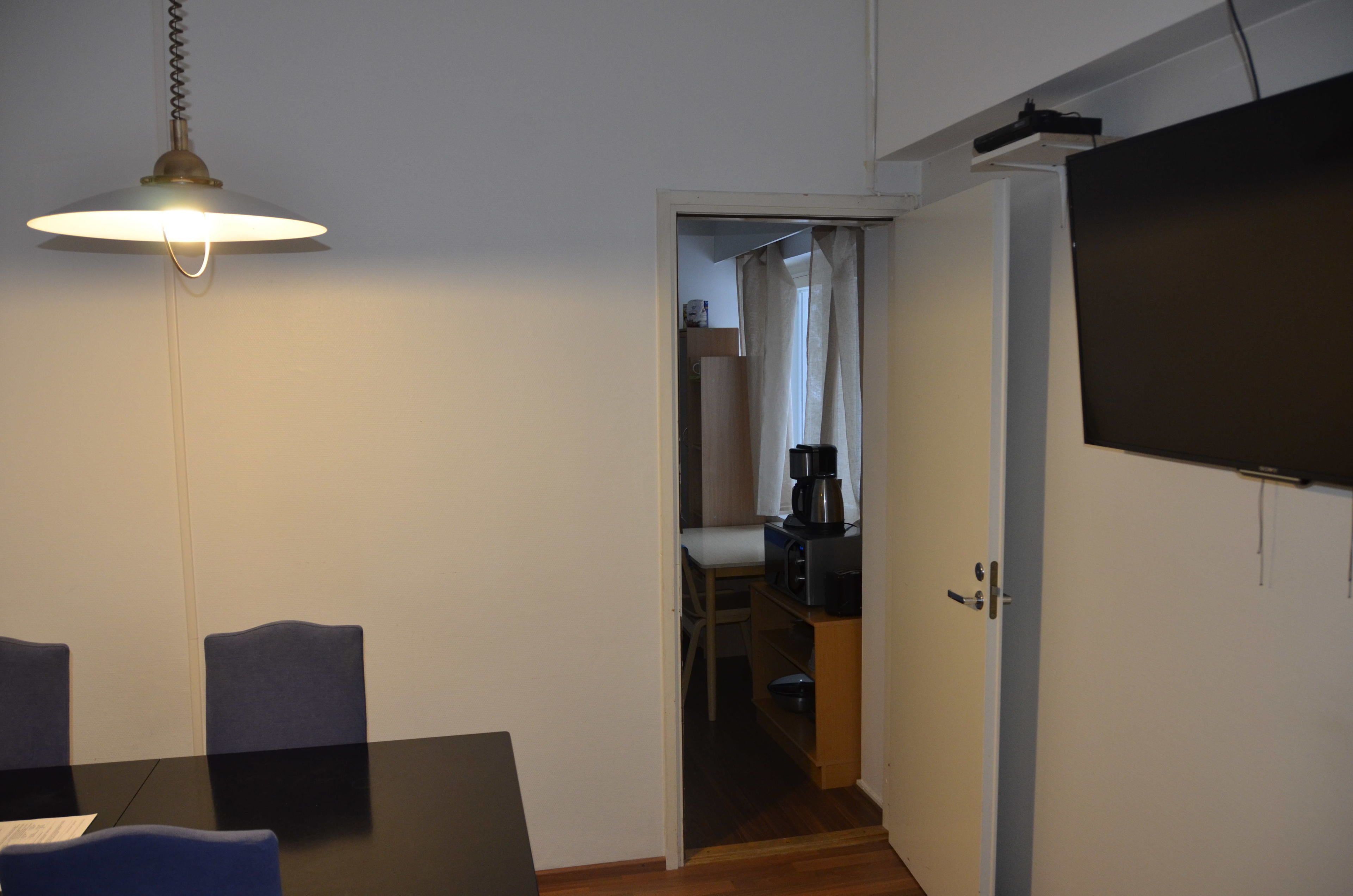 Een kamer met een eettafel, stoelen, een hanglamp, een televisie en een deuropening naar een keuken.