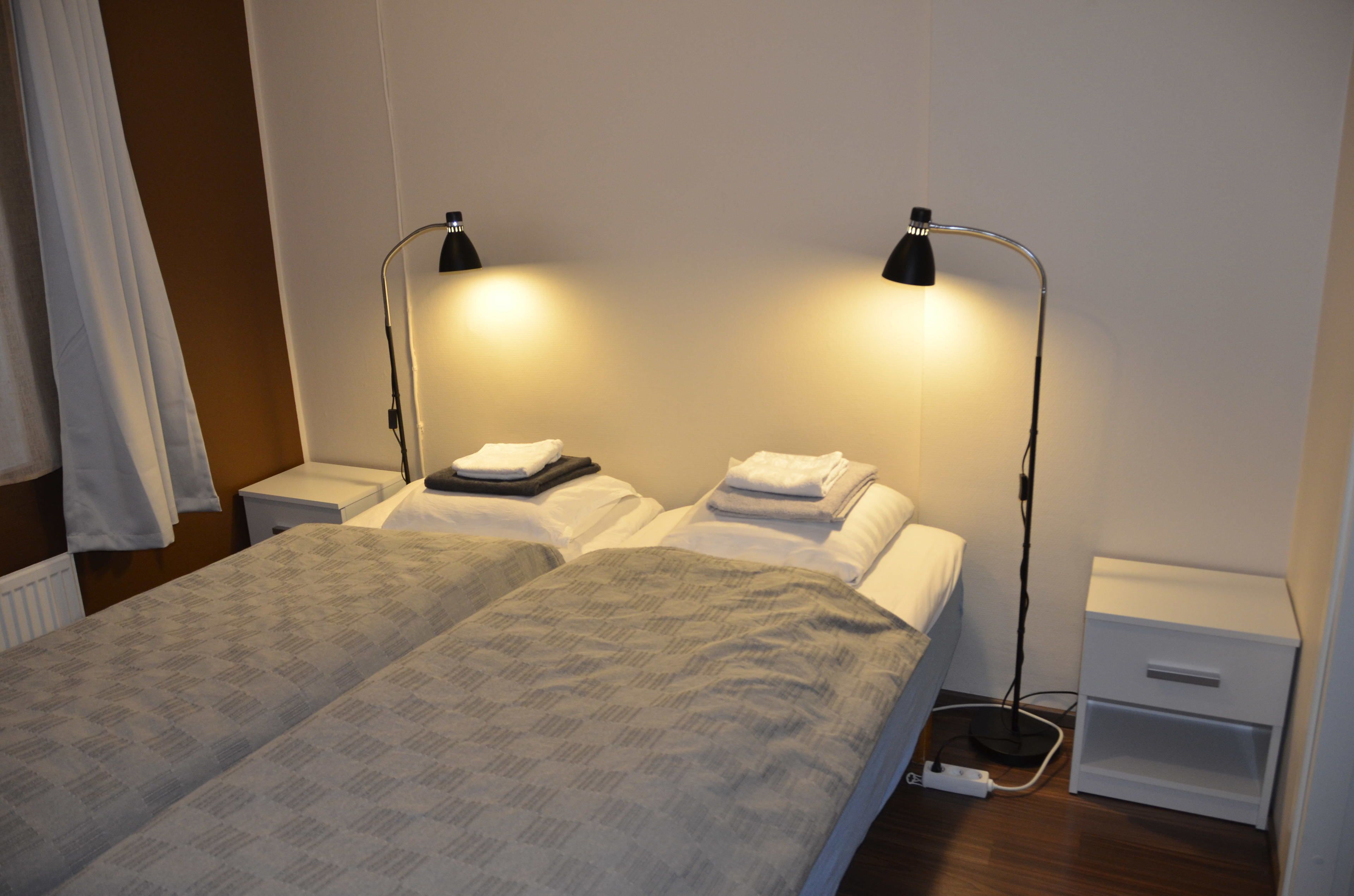 Camera d'albergo con due letti, ciascuno con un cuscino e un asciugamano piegato. Sono presenti due comodini, uno con una lampada e l'altro con una presa di corrente.