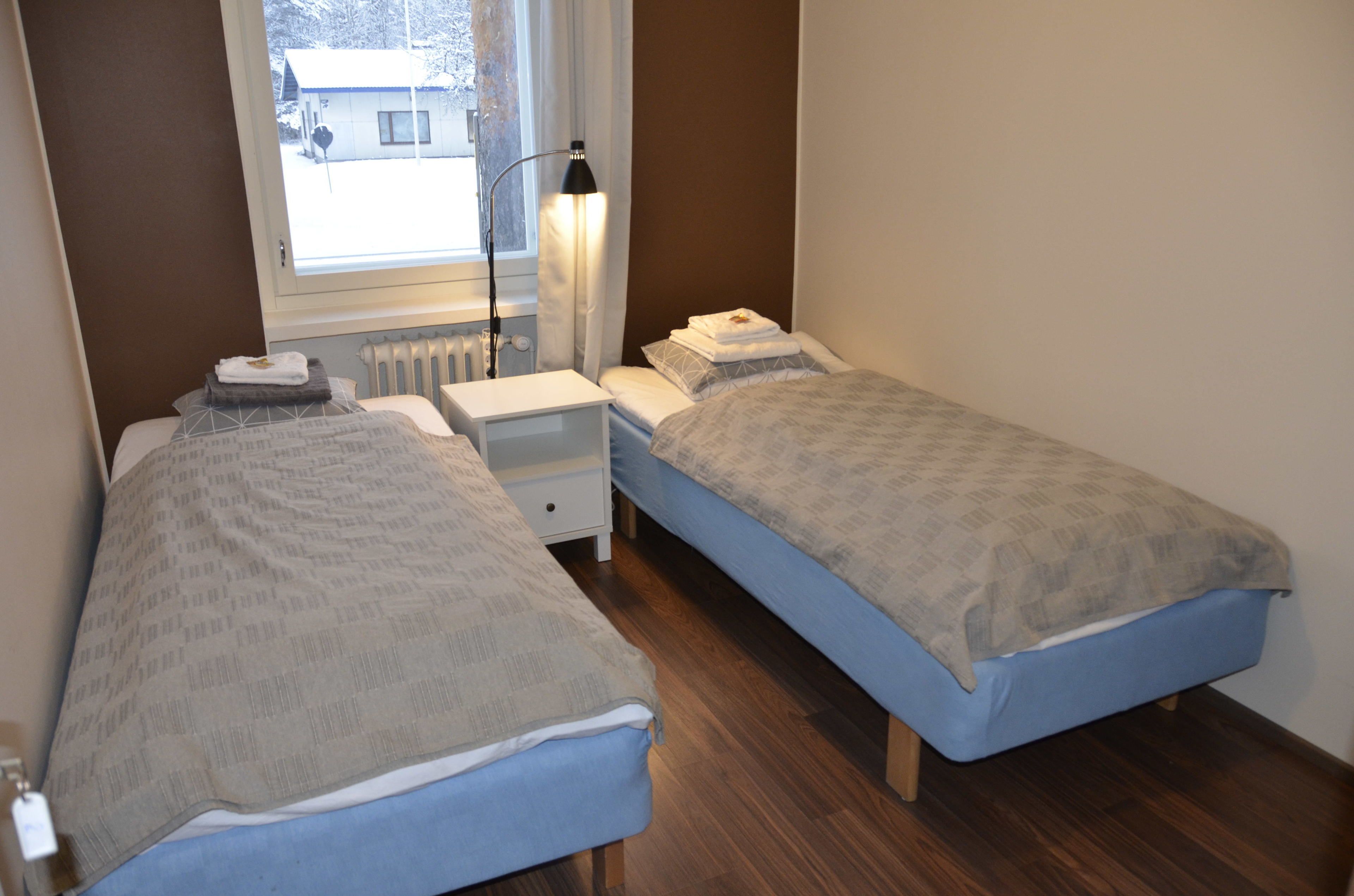 A bedroom with two beds, a nightstand, a lamp, and a window with a view of a building outside.