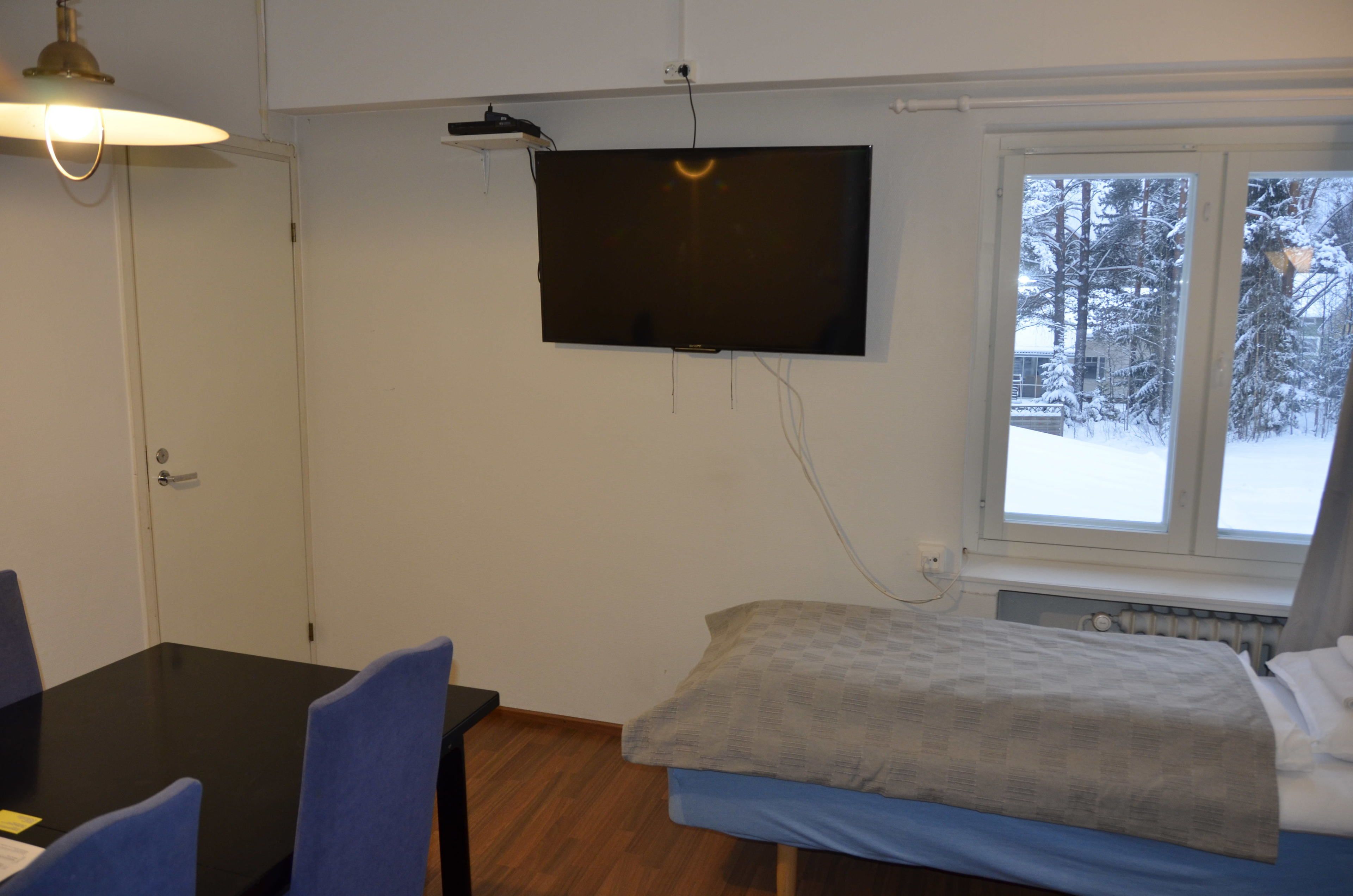 Una habitación con cama, una mesa con sillas, un televisor fijado en la pared y una ventana con vistas a árboles cubiertos de nieve.