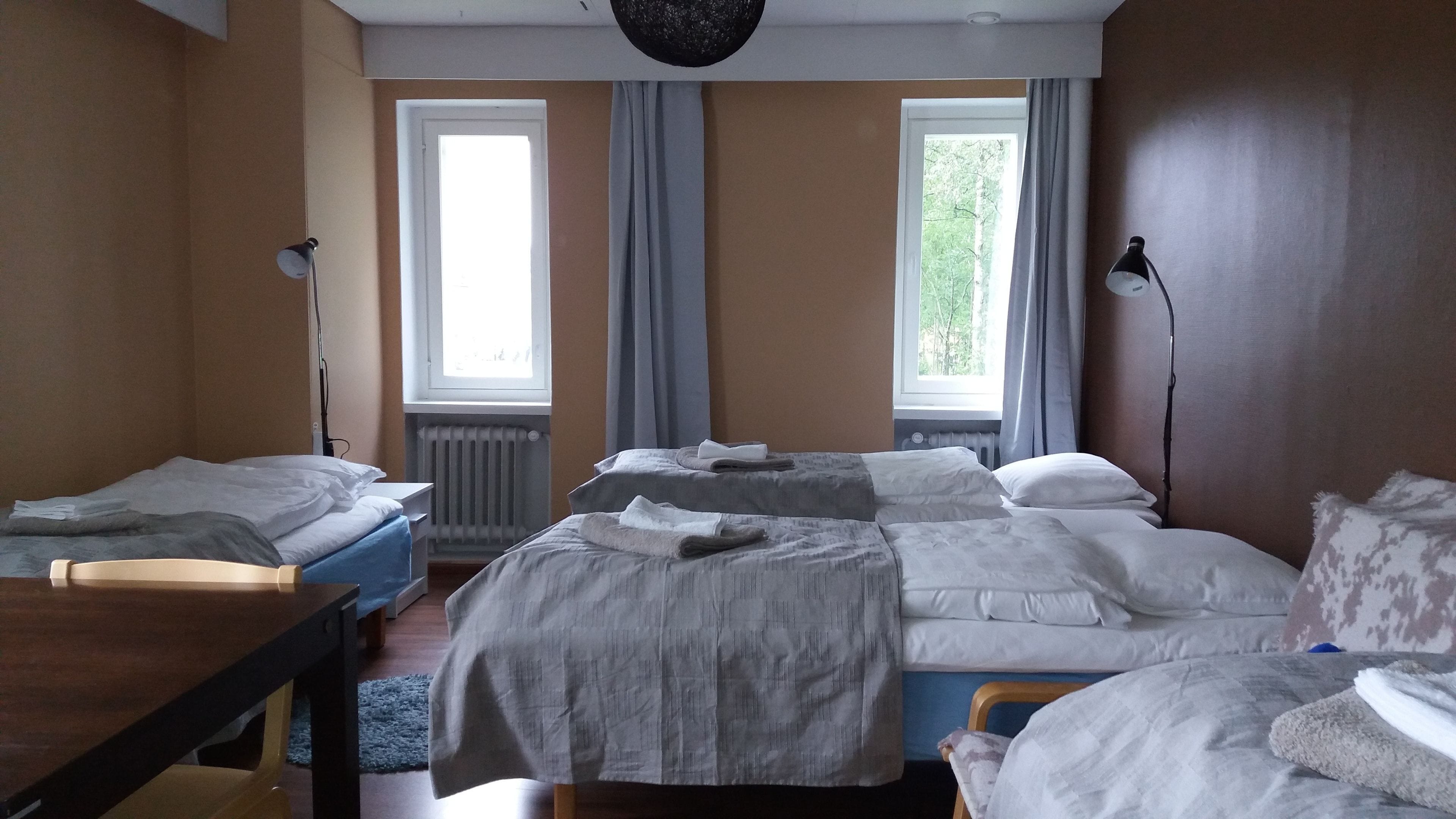 Habitación de hotel con dos camas, un escritorio, una silla, un radiador y dos ventanas con cortinas.