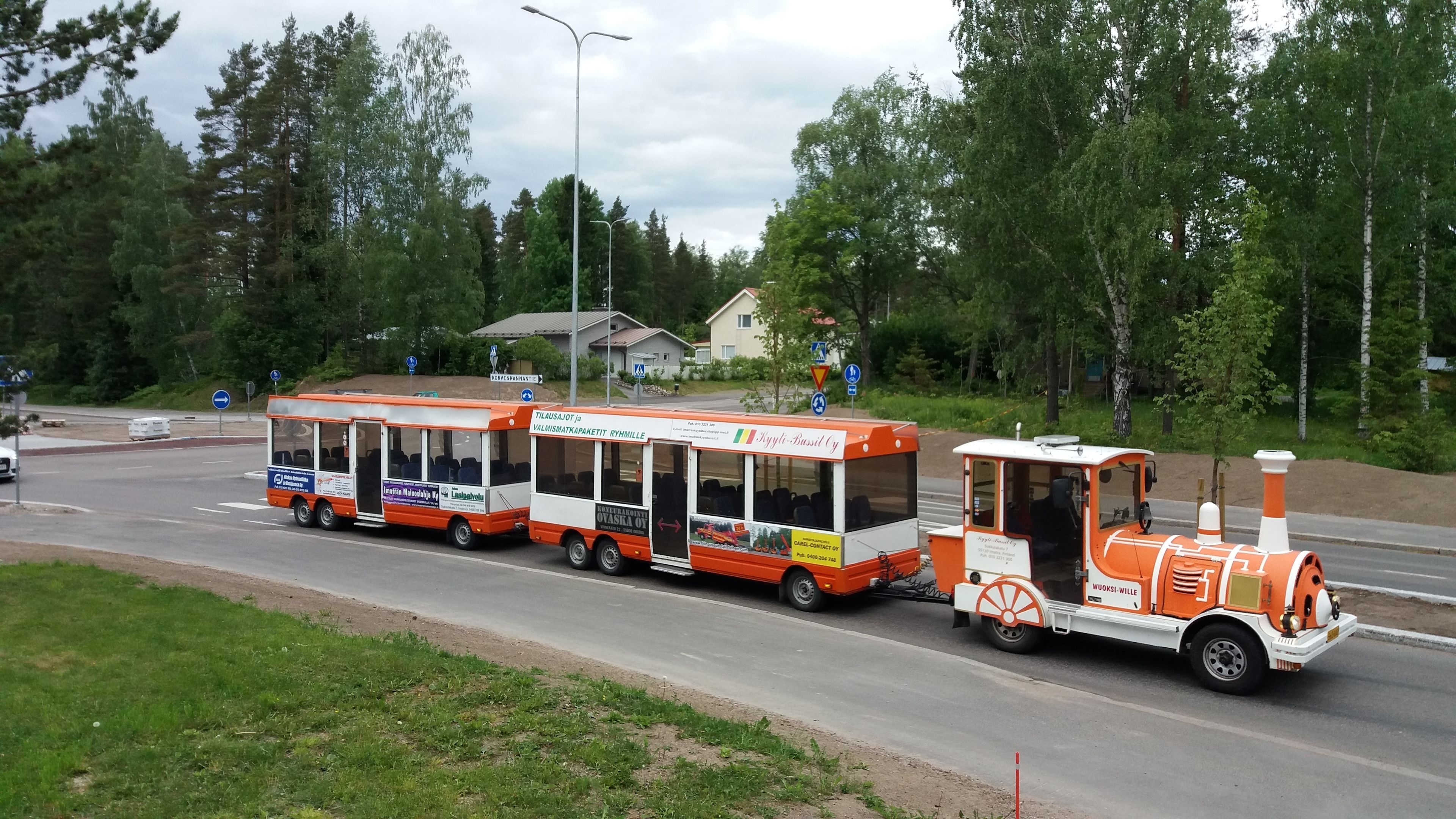 Un train à voie étroite, orné de publicités sur ses flancs, circule sur une route bordée d’arbres, avec une maison visible au loin.