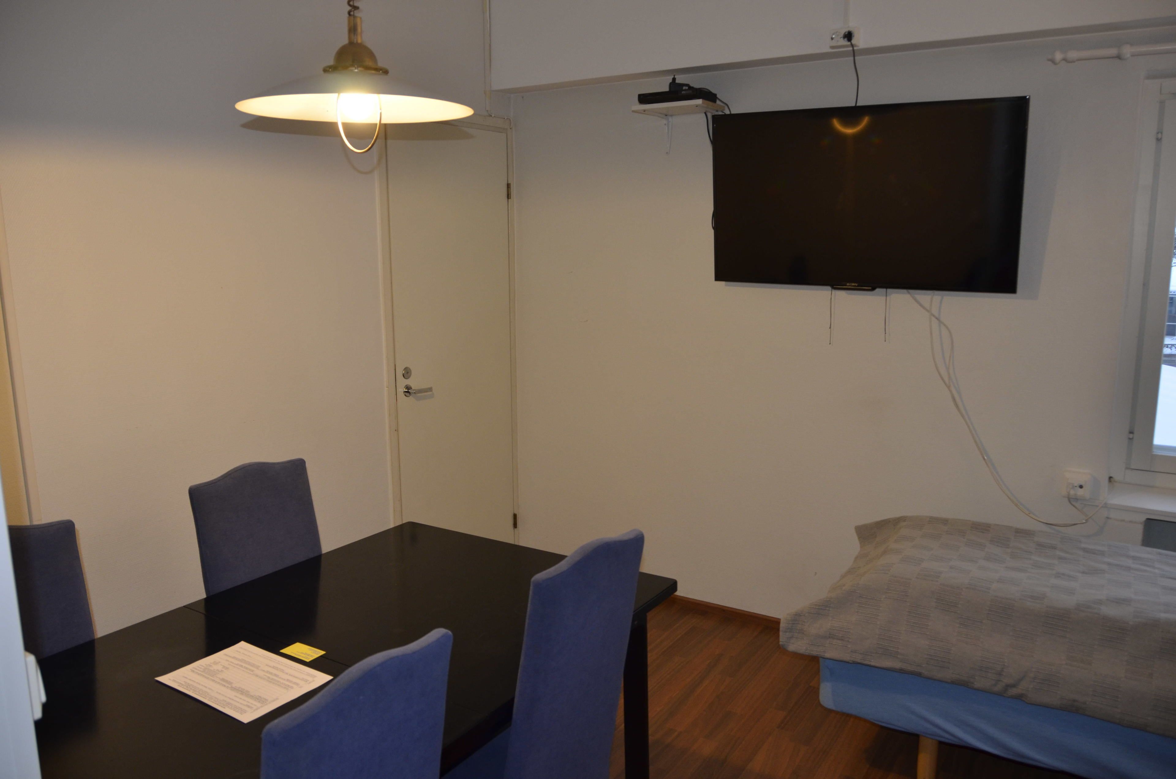 Una habitación con cama, televisión montada en la pared, una mesa con sillas y una lámpara.