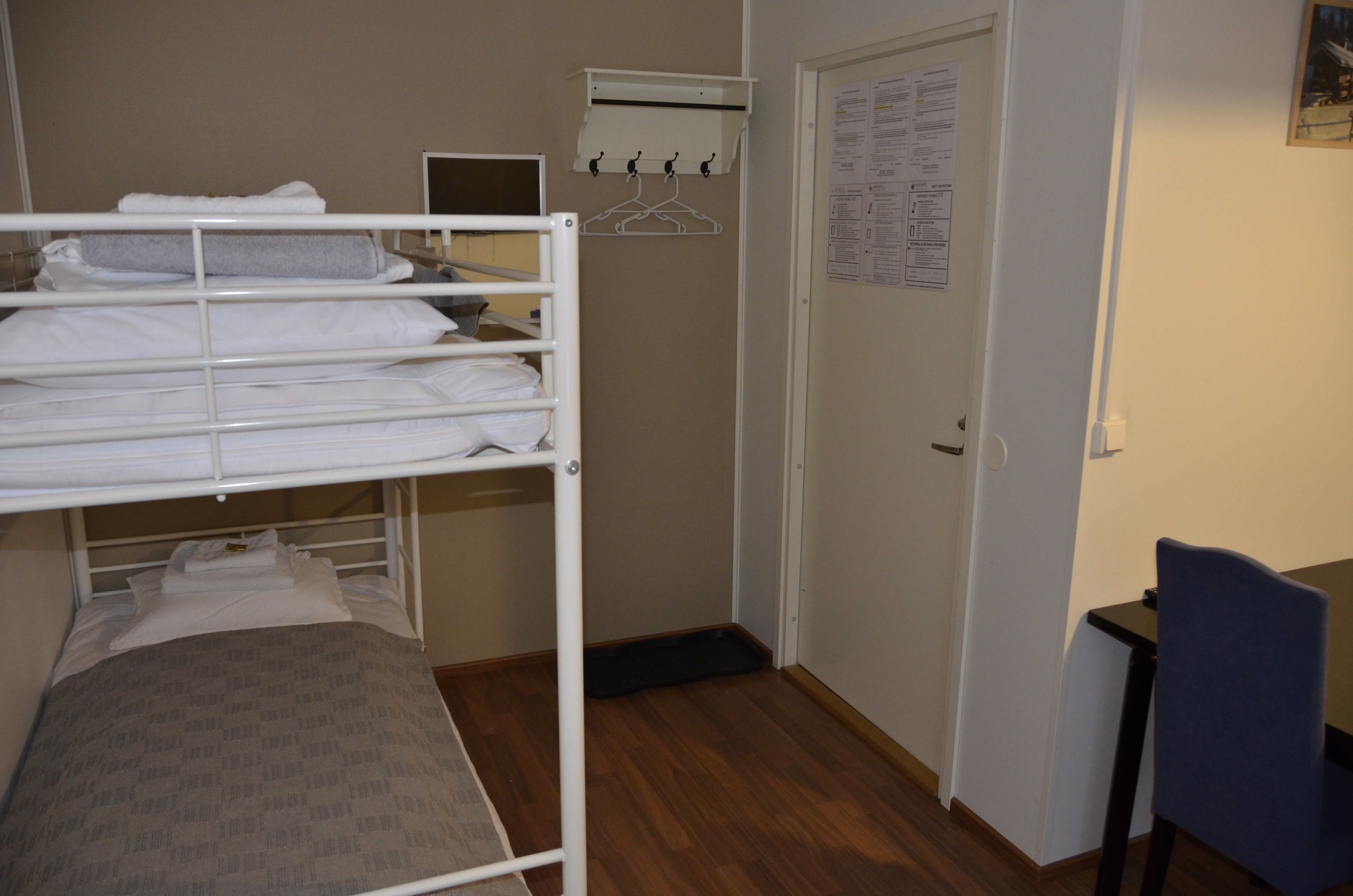 Una cama individual con ropa de cama blanca, un escritorio con silla, una puerta con un aviso y un perchero con perchas.
