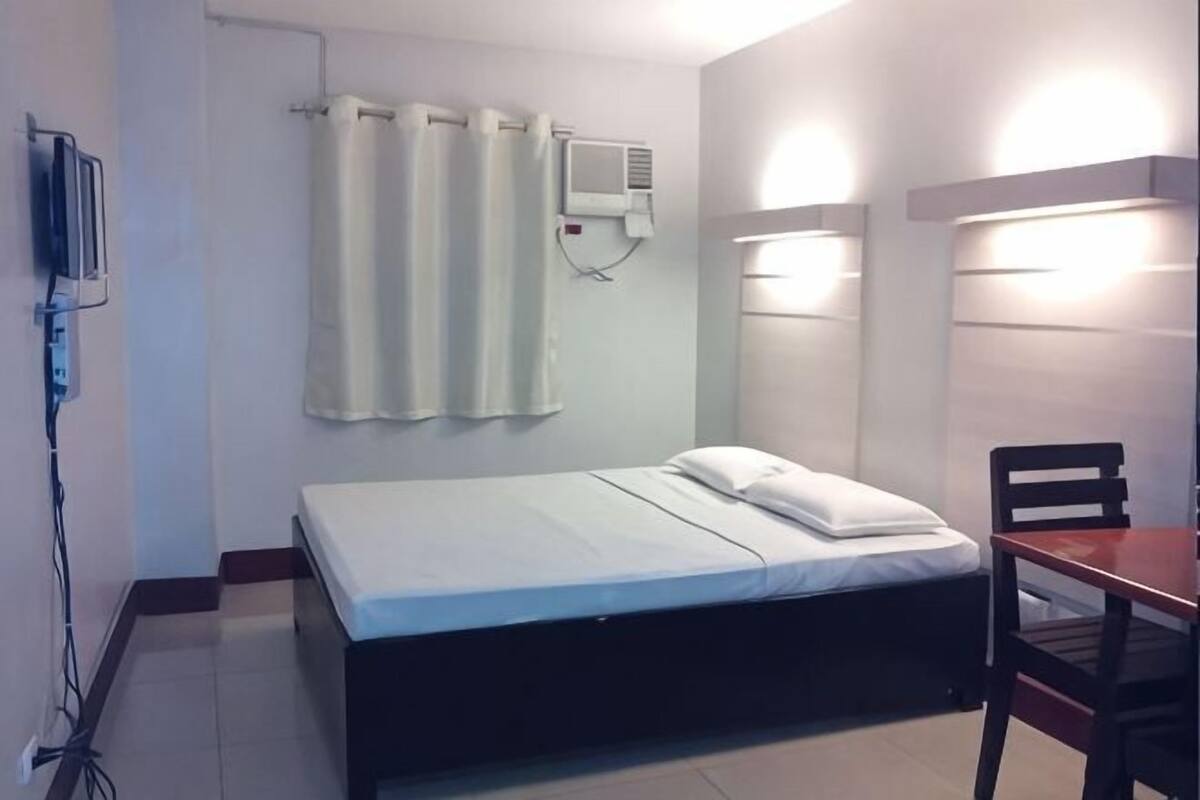 A single bed with a white mattress and pillows, a bedside table, a chair, a wall-mounted air conditioner, and a curtain.