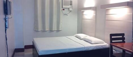 Una cama individual con colchón y almohadas blancos, una mesita de noche, una silla, un aire acondicionado de pared y una cortina.