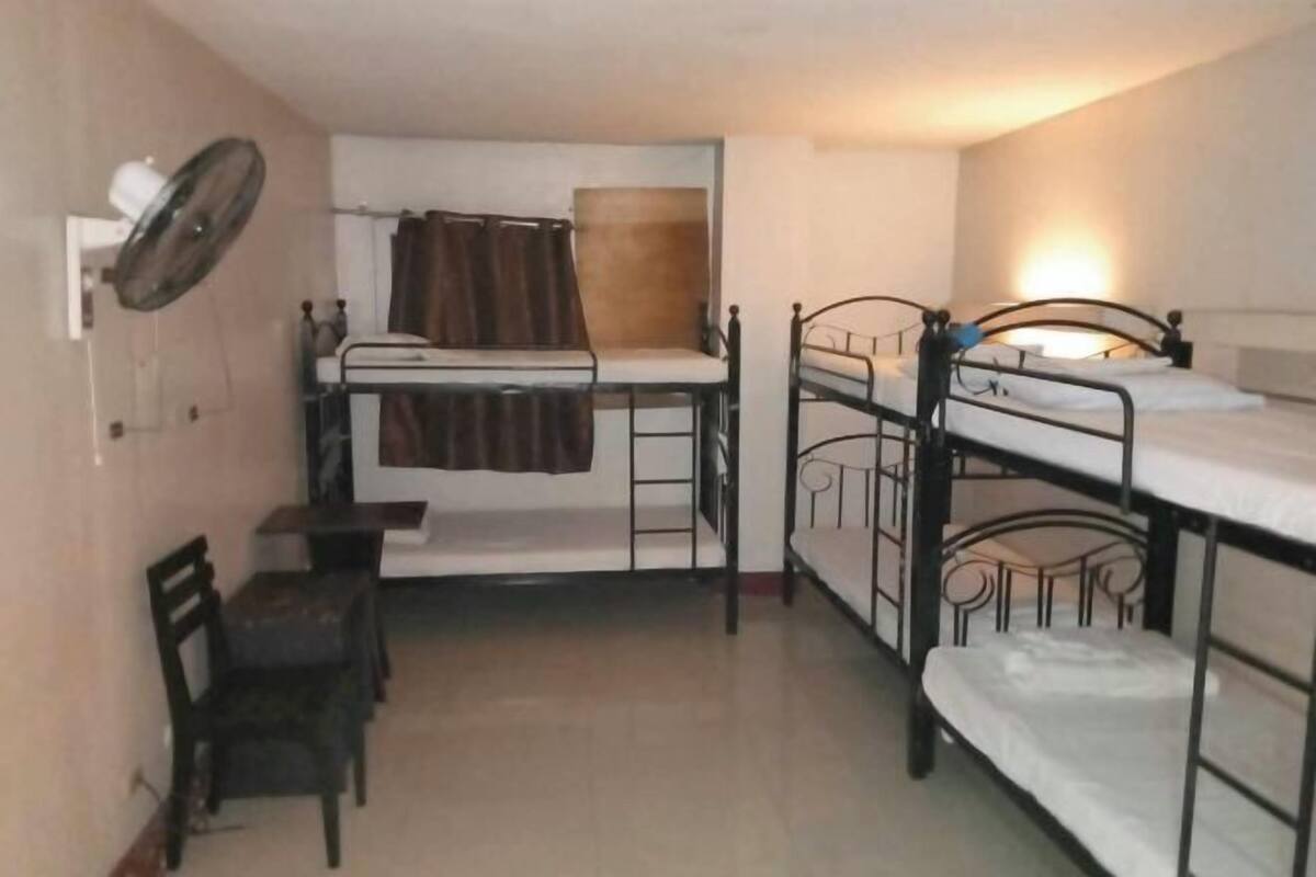 A dormitory room with bunk beds, a desk, a chair, and a fan.