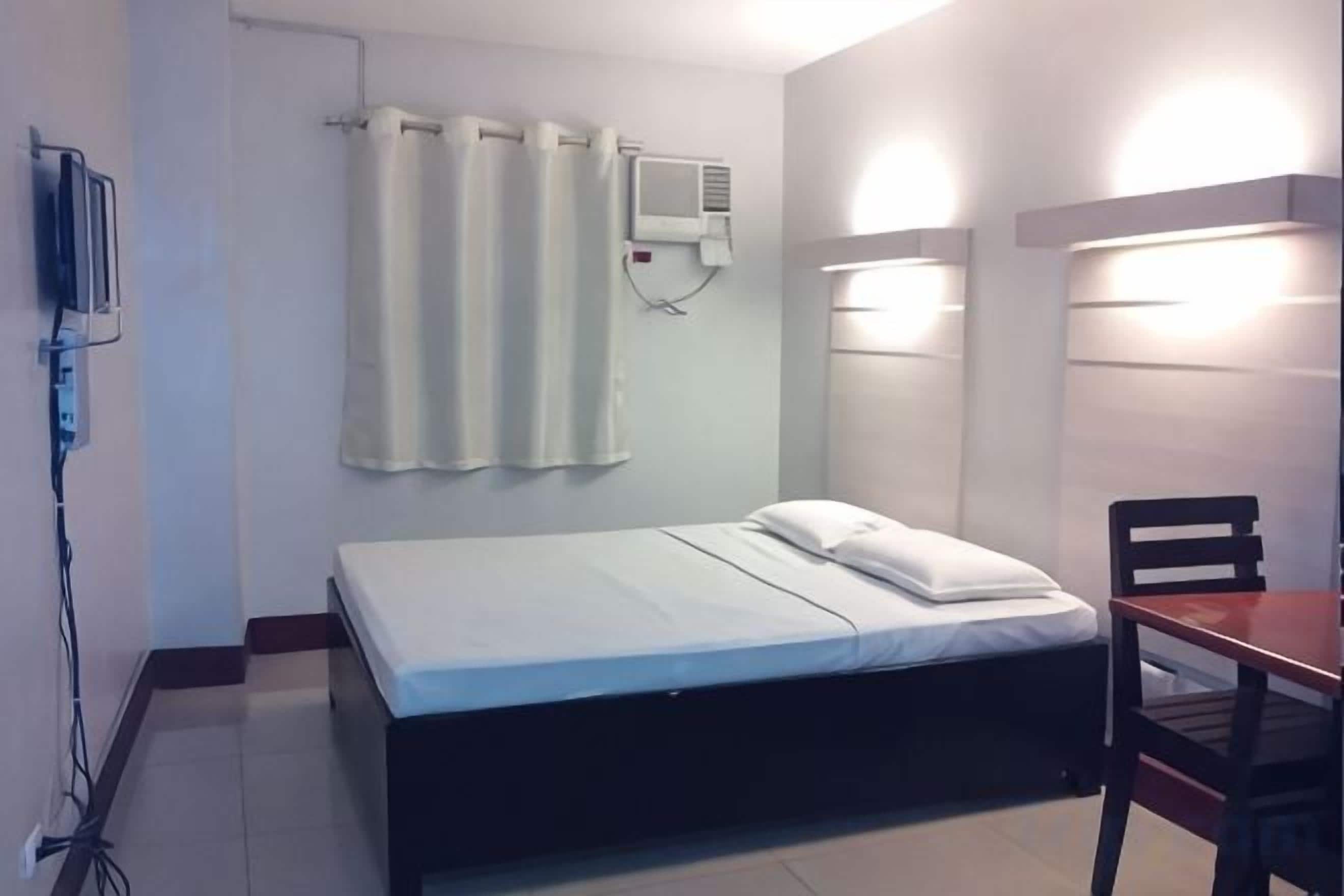 Una cama individual con colchón y almohadas blancas, una mesita de noche, una silla, un aire acondicionado de pared y una cortina.