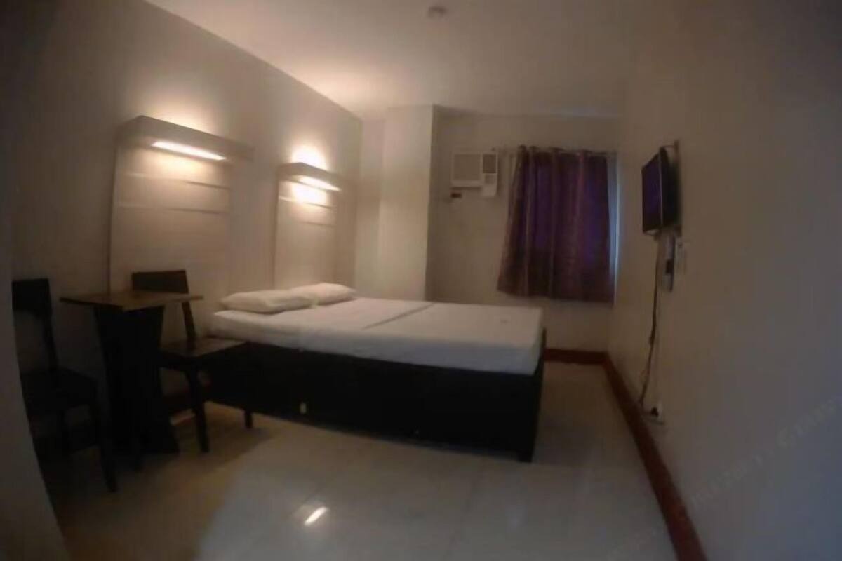 A small, modern room with a bed, a desk, and a chair. There are wall-mounted lights and a flat-screen TV.
