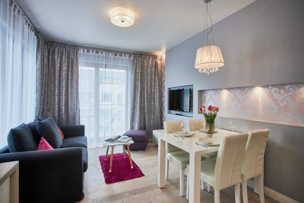 Una sala de estar moderna con un sofá, una mesa de comedor con sillas, un televisor montado en la pared y una ventana con cortinas.