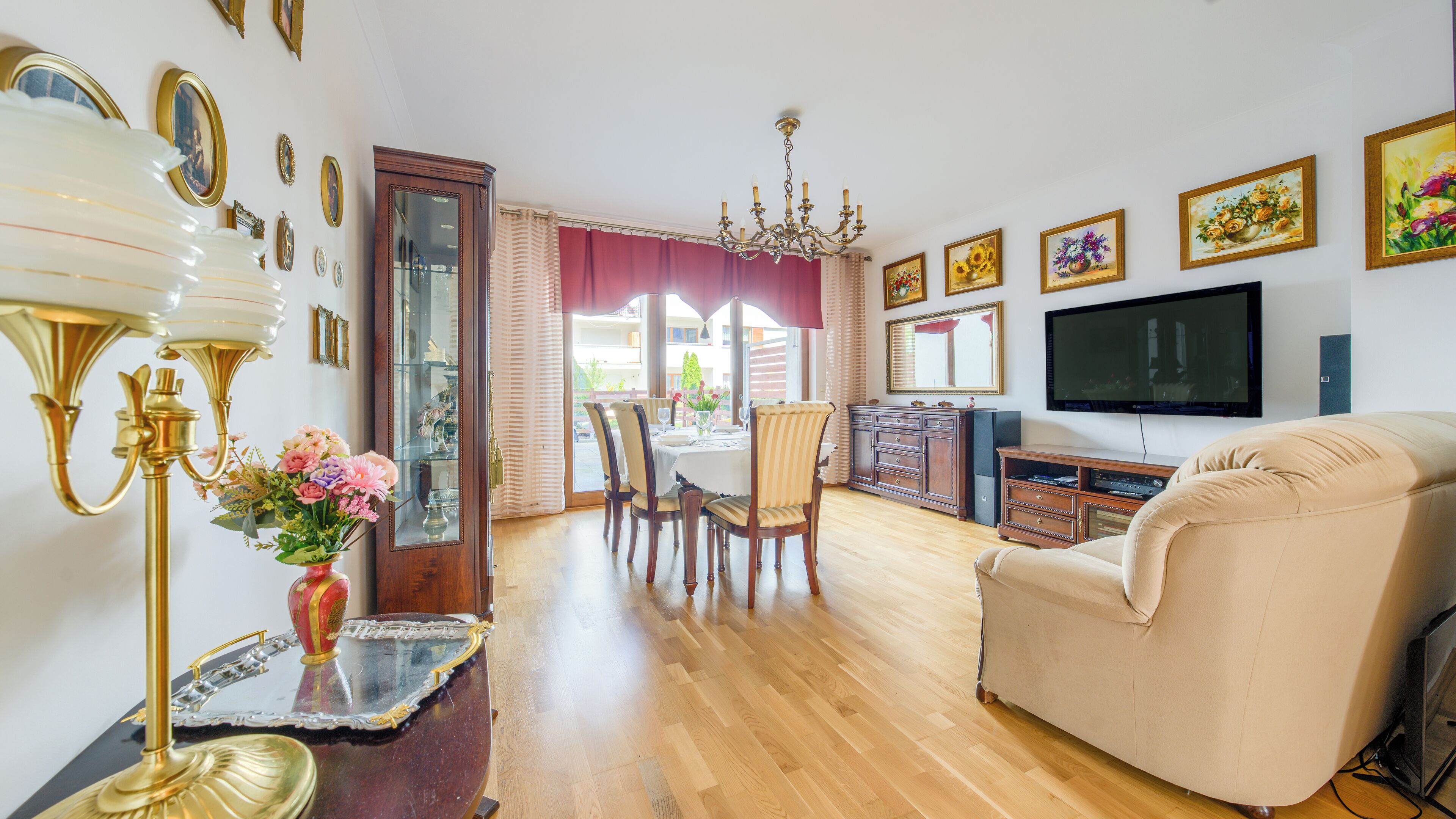 Een woonkamer met een bank, televisie, houten kast, eettafel met stoelen en een kroonluchter.