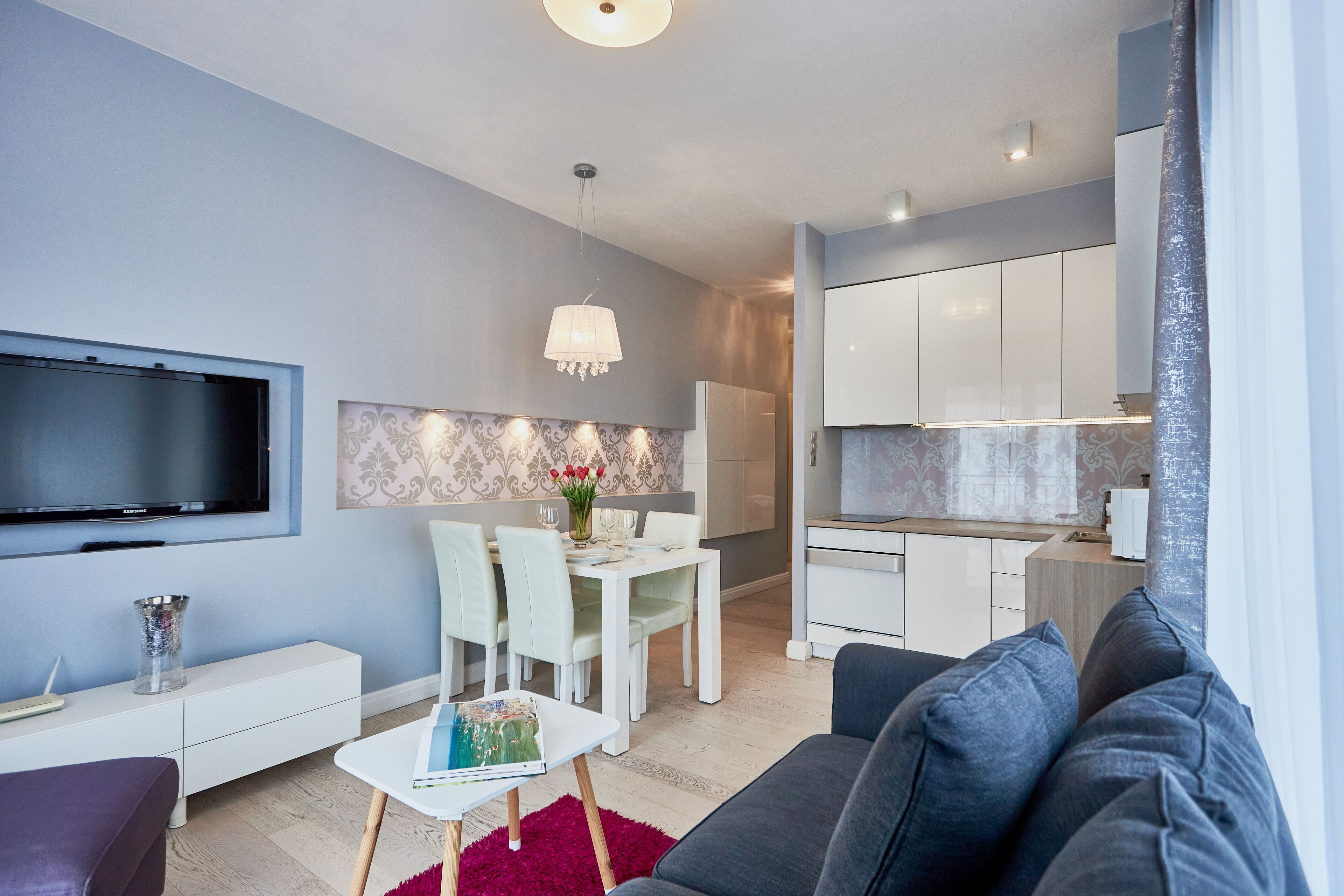 A modern living room with a flat-screen TV, a dining area with a white table and chairs, and a kitchen with white cabinets.