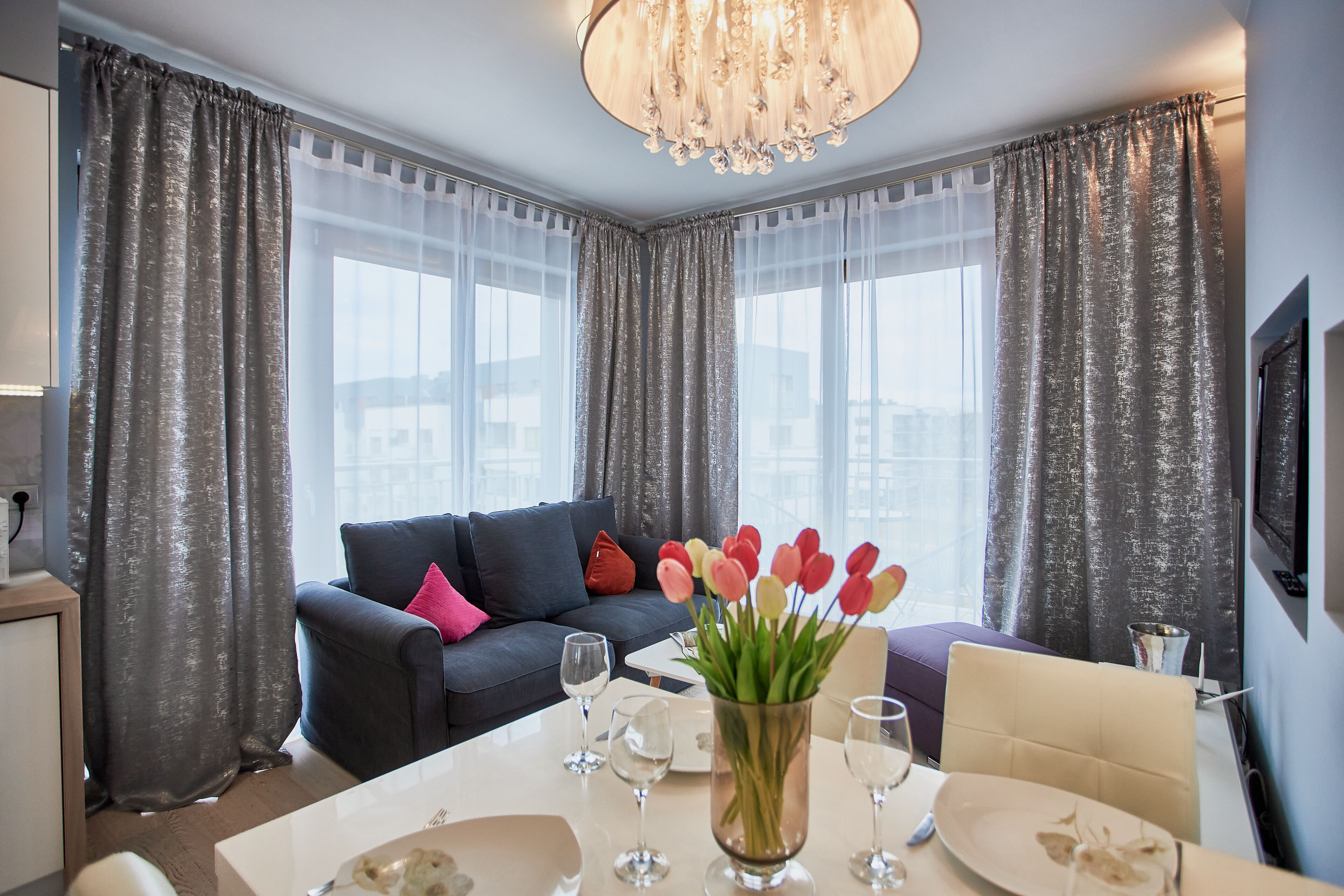 A modern living room with a dark sofa, a dining table set with glasses, and a vase of tulips.