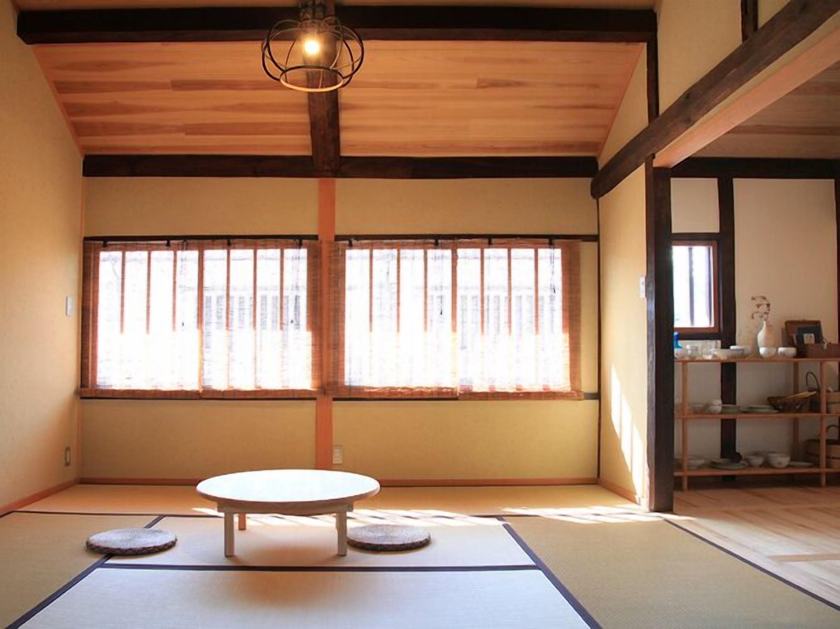 A tatami mat room with a tatami mat table, two cushions, a hanging light fixture, and a window with vertical blinds.