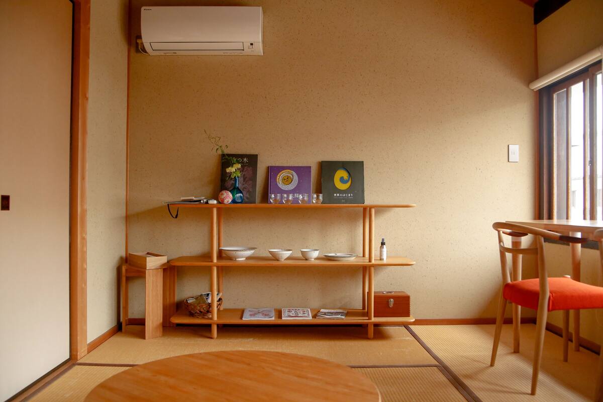 Habitació amb paviment de tatami, taula de fusta, cadira, prestatge amb bols i aire condicionat fixat a la paret.